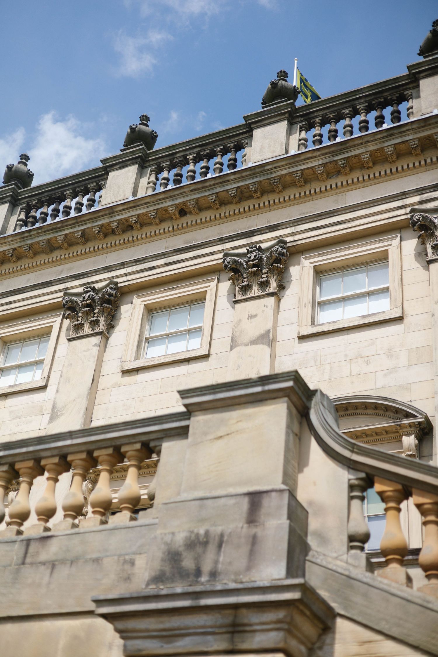 Grand neoclassical stone building facade with ornate balustrades, carved corbels, and sash windows under a blue sky.
