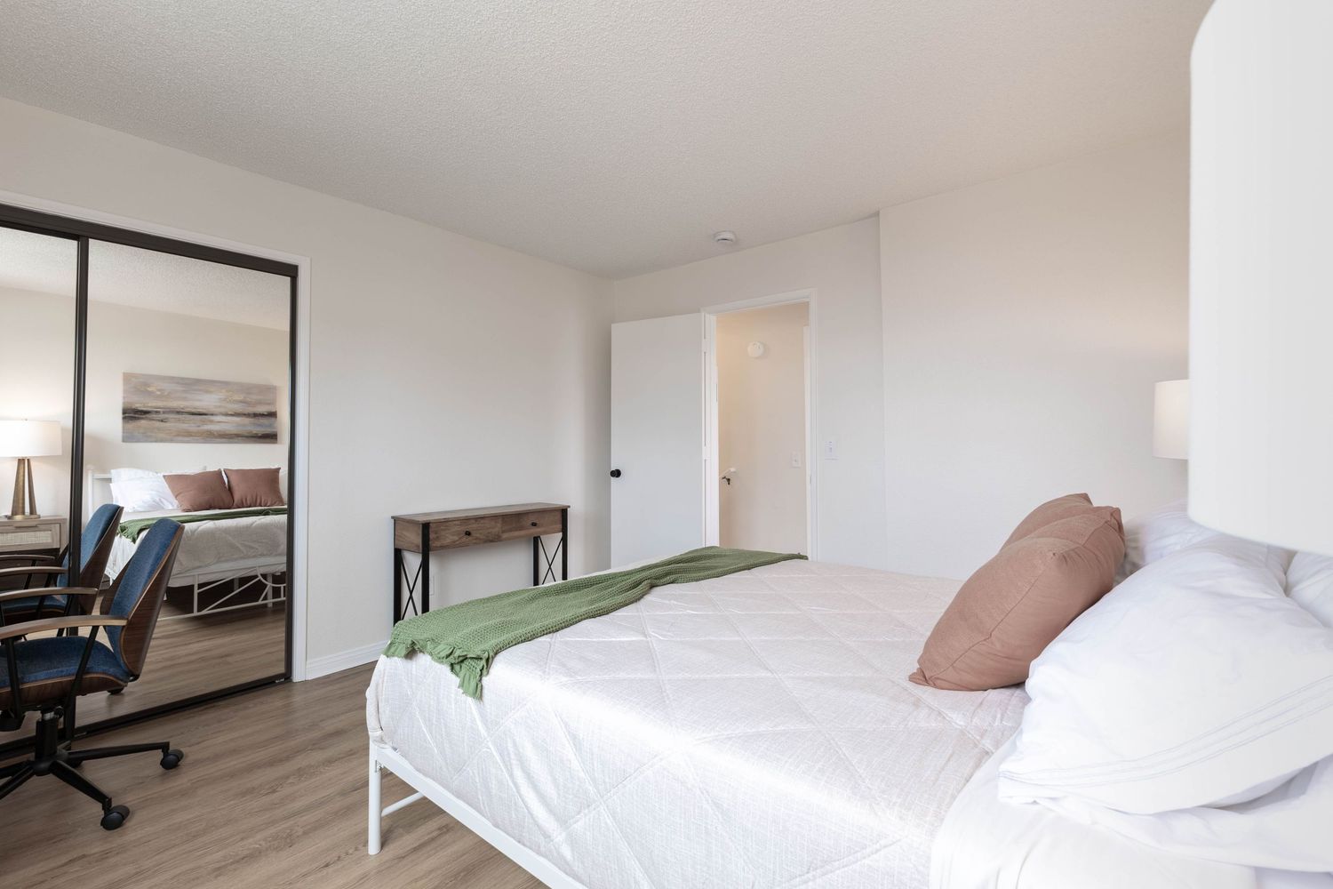 Modern bedroom with white bedding, mirrored closet doors, hardwood floors, and a desk area with office chair.