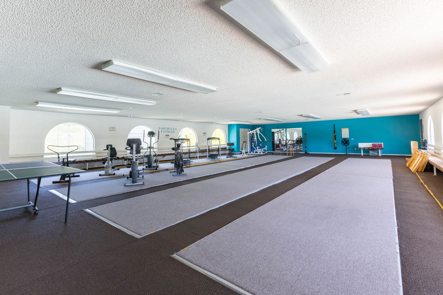 Spacious fitness center with cardio equipment, wall mirrors, carpet, and a ping pong table in a bright room with teal accent wall.
