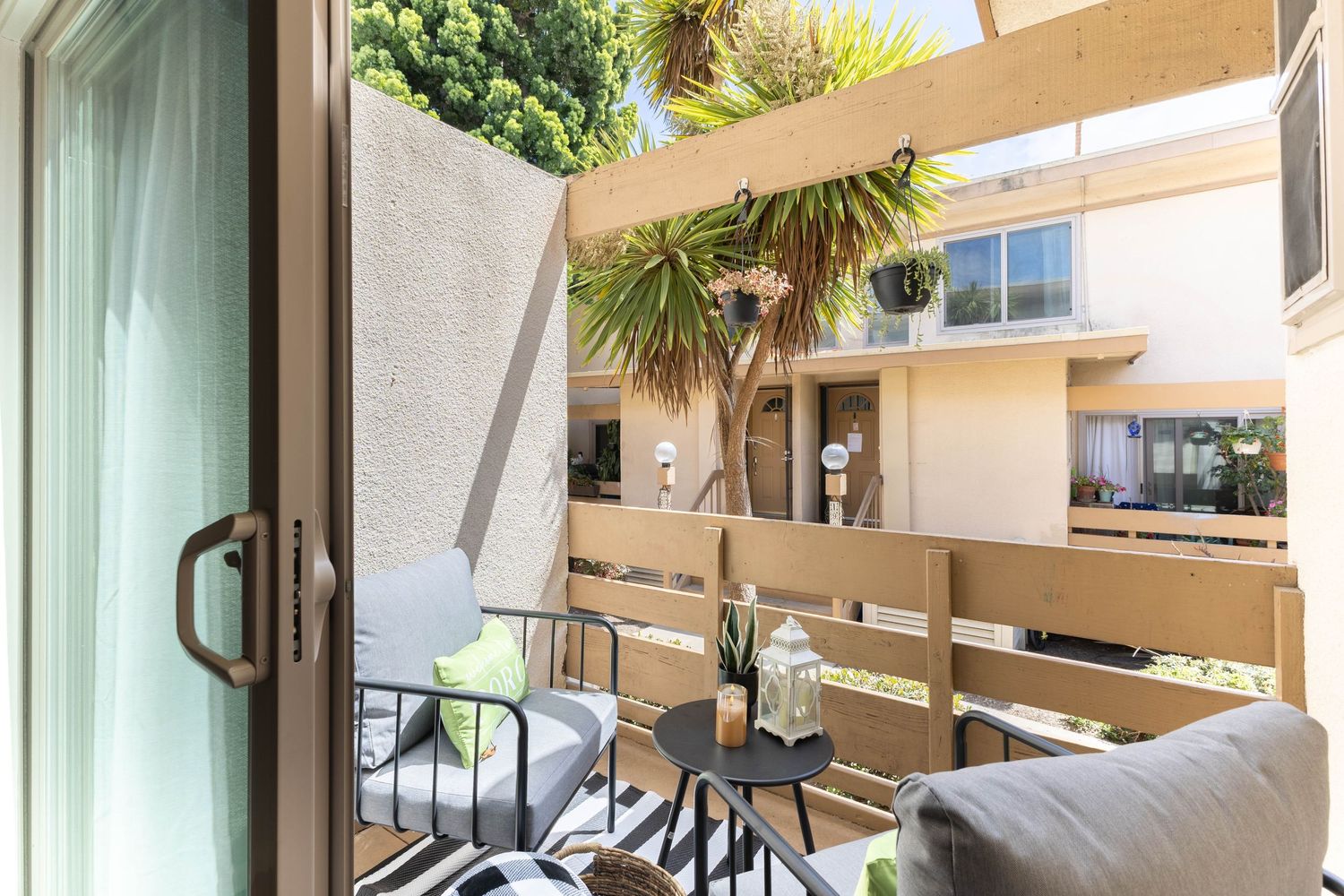 Cozy apartment balcony with patio chairs, small table, wood railings, and lush tropical plants in a sunny courtyard.