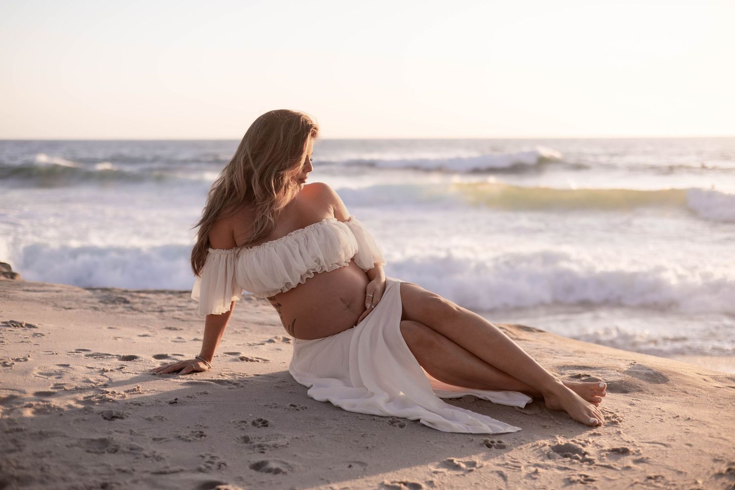 Pregnant woman in white dress sitting on sandy beach with ocean waves at sunset.