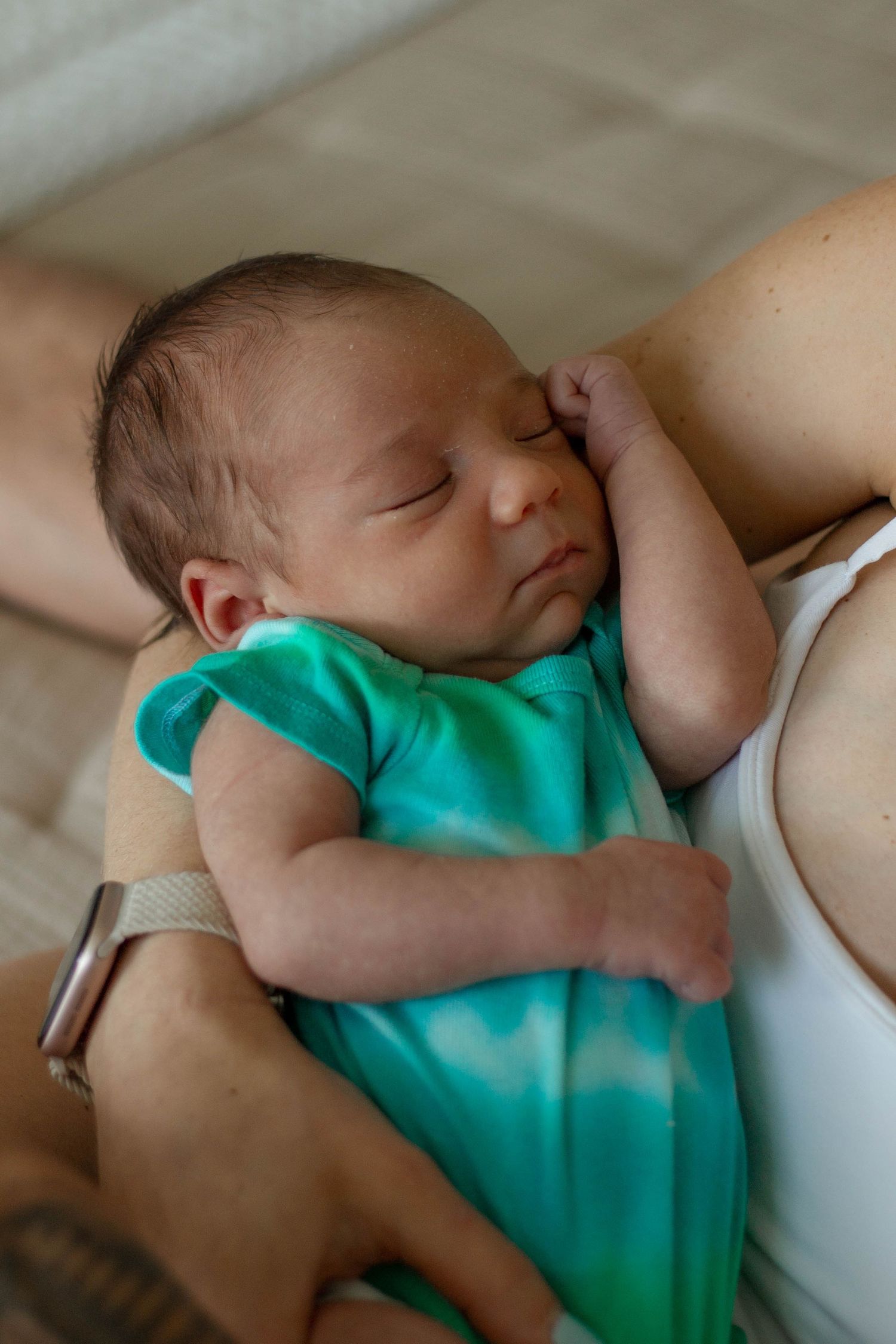 Peaceful newborn baby sleeping in teal onesie cradled in mother's arms.