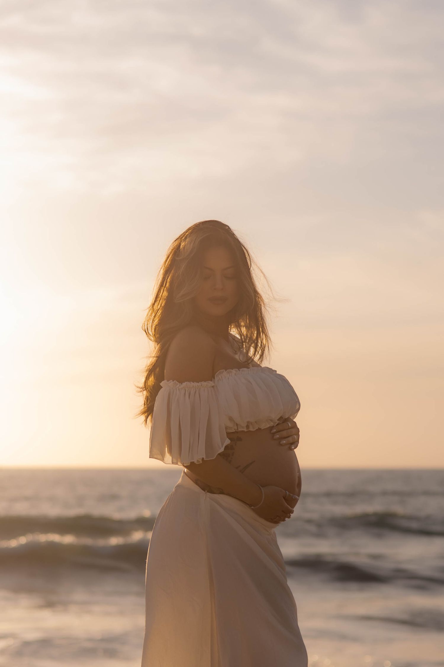 Pregnant woman in white off-shoulder dress cradling bump at golden ocean sunset.