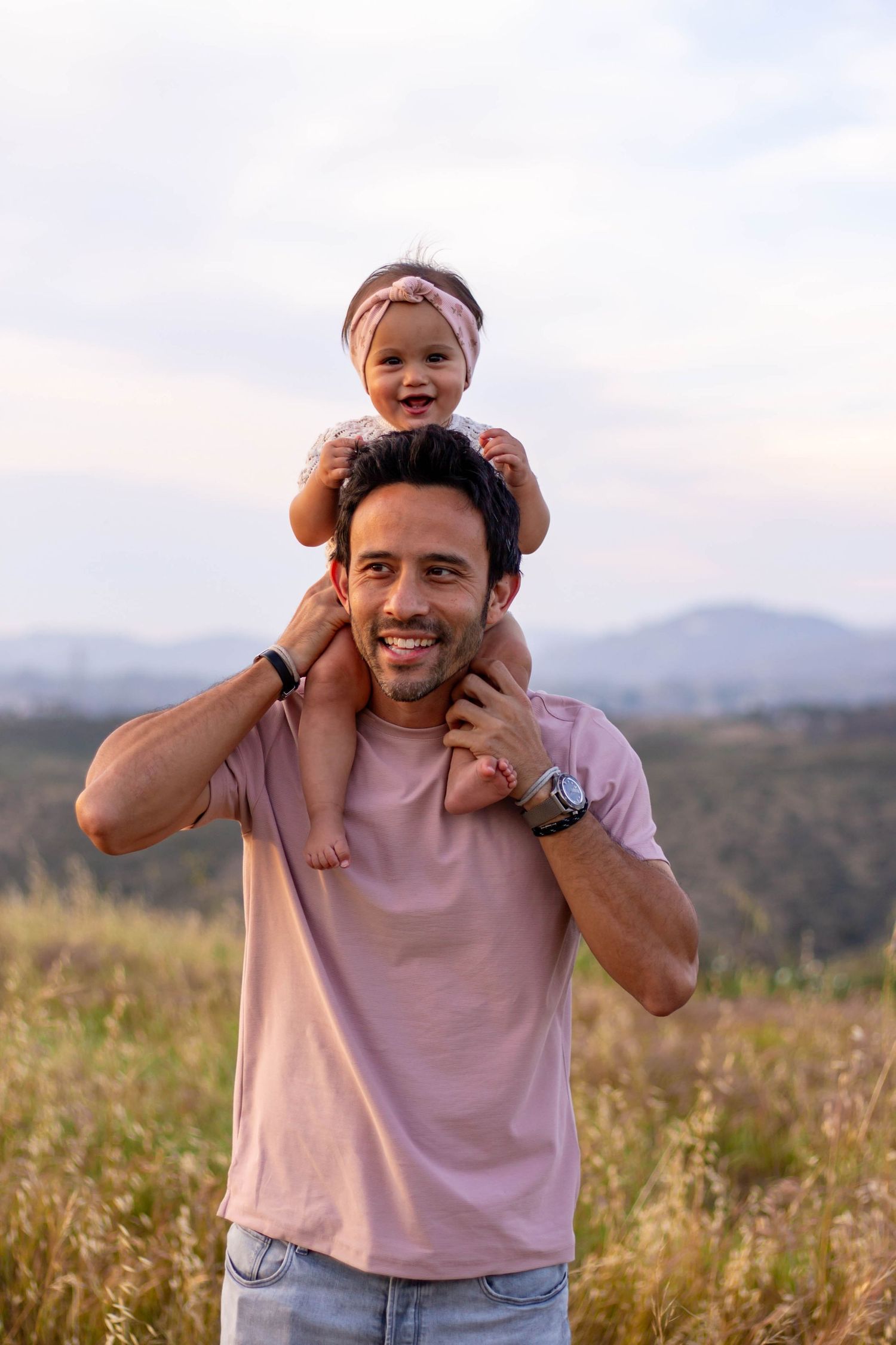 Smiling father carrying laughing baby girl on shoulders in scenic hilltop meadow at dusk.