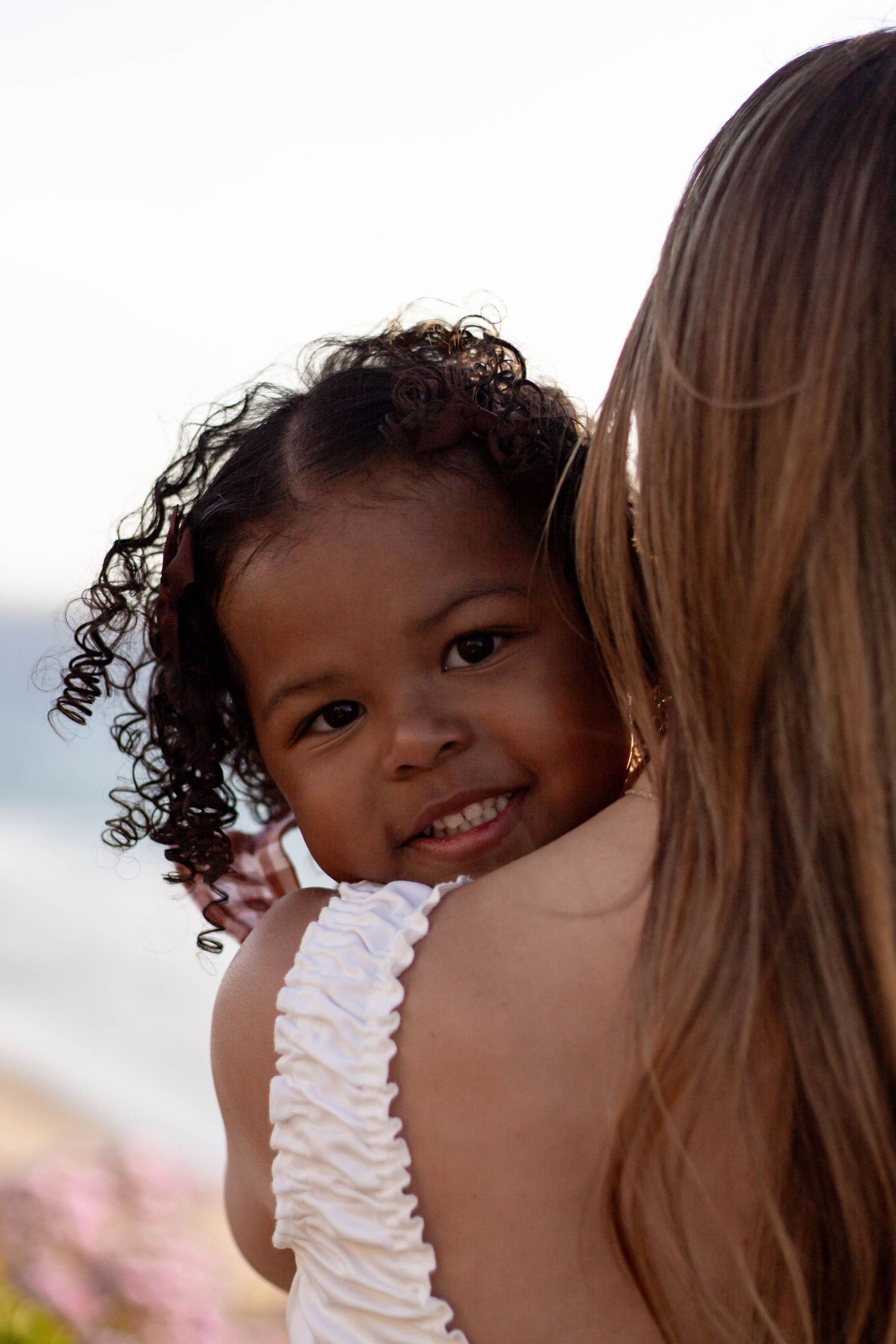 Happy curly-haired toddler girl smiling over the shoulder of a blonde woman outdoors.