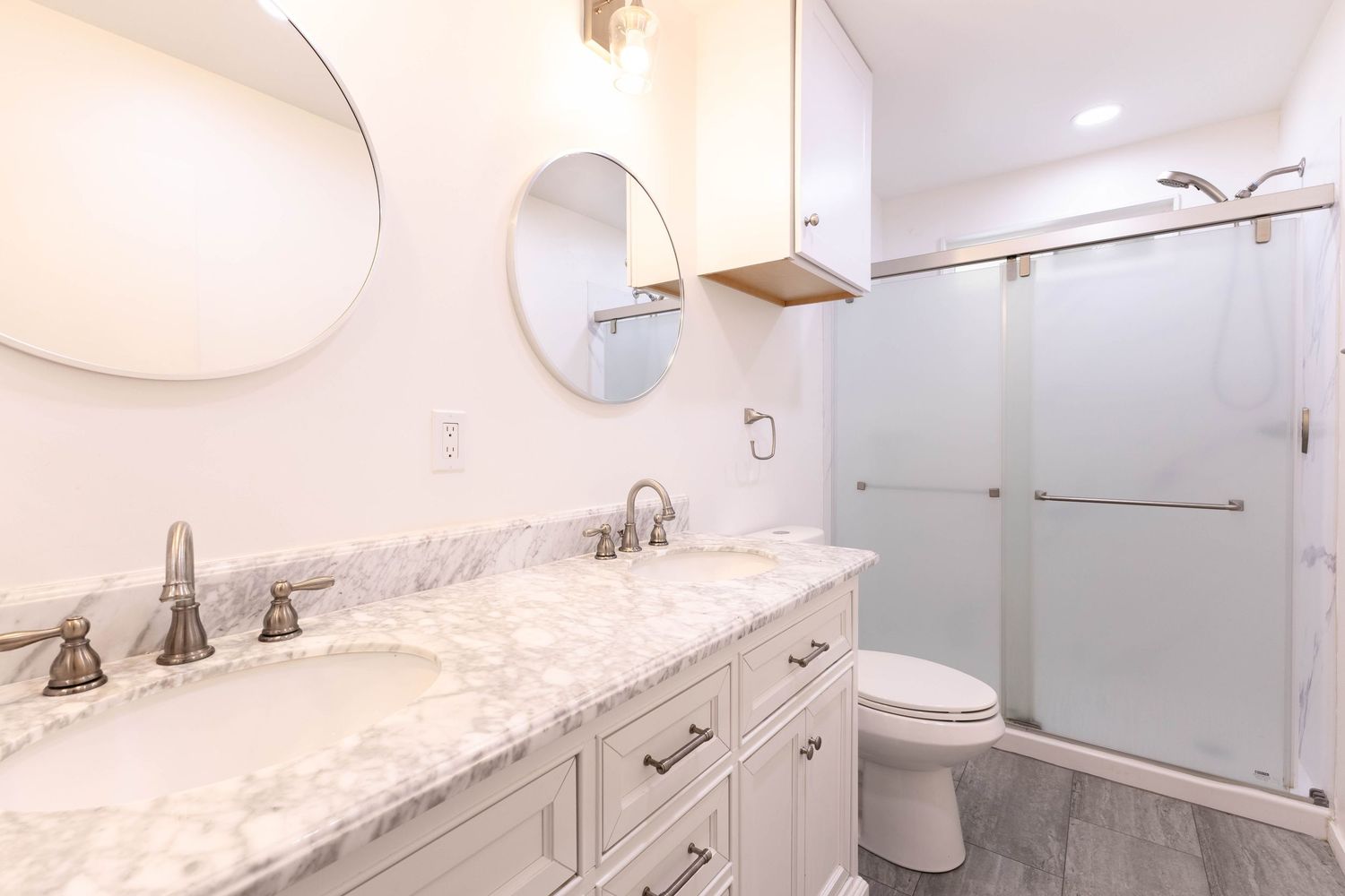 Modern white bathroom with marble vanity, dual sinks, round mirrors, glass shower, and wood-look tile flooring.