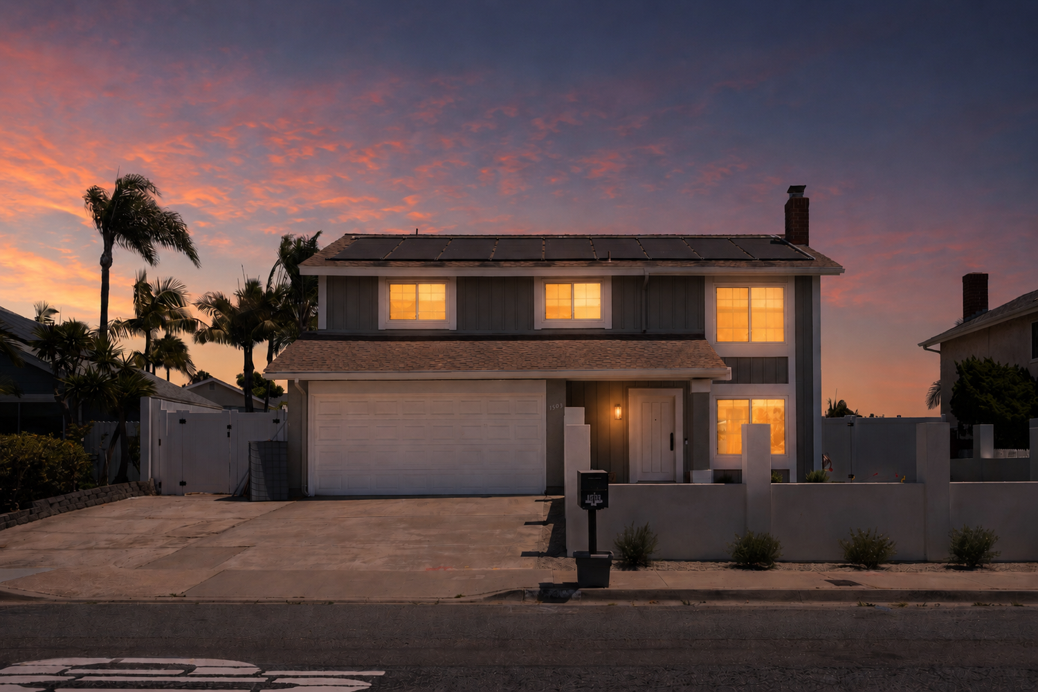 Two-story suburban home with glowing windows against a vibrant sunset sky, featuring a two-car garage and palm trees.