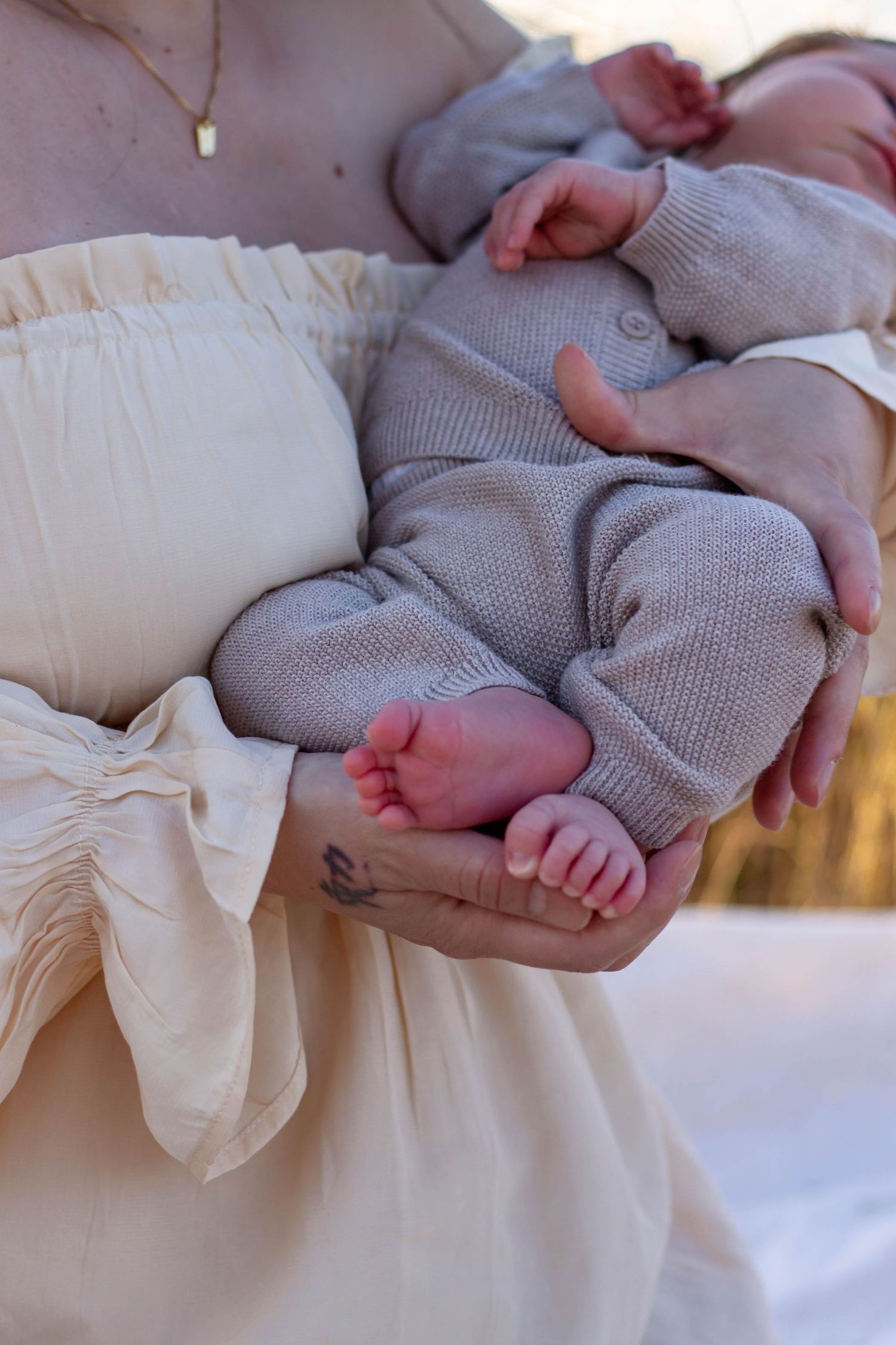Mother in cream dress cradling sleeping newborn baby in knit outfit, showing tiny feet and hands.