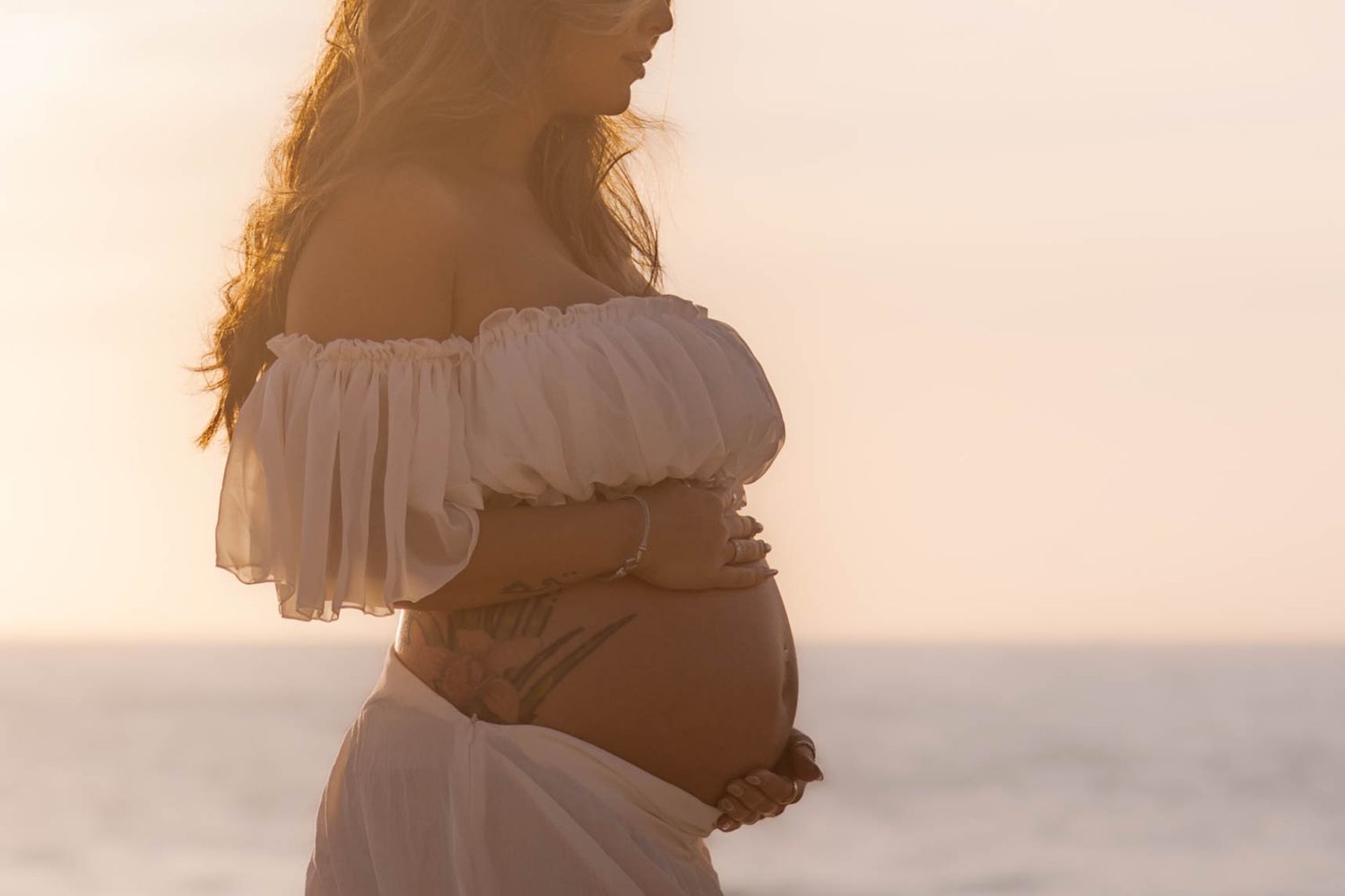 Silhouette of pregnant woman in white dress cradling bump against warm golden sunset beach.