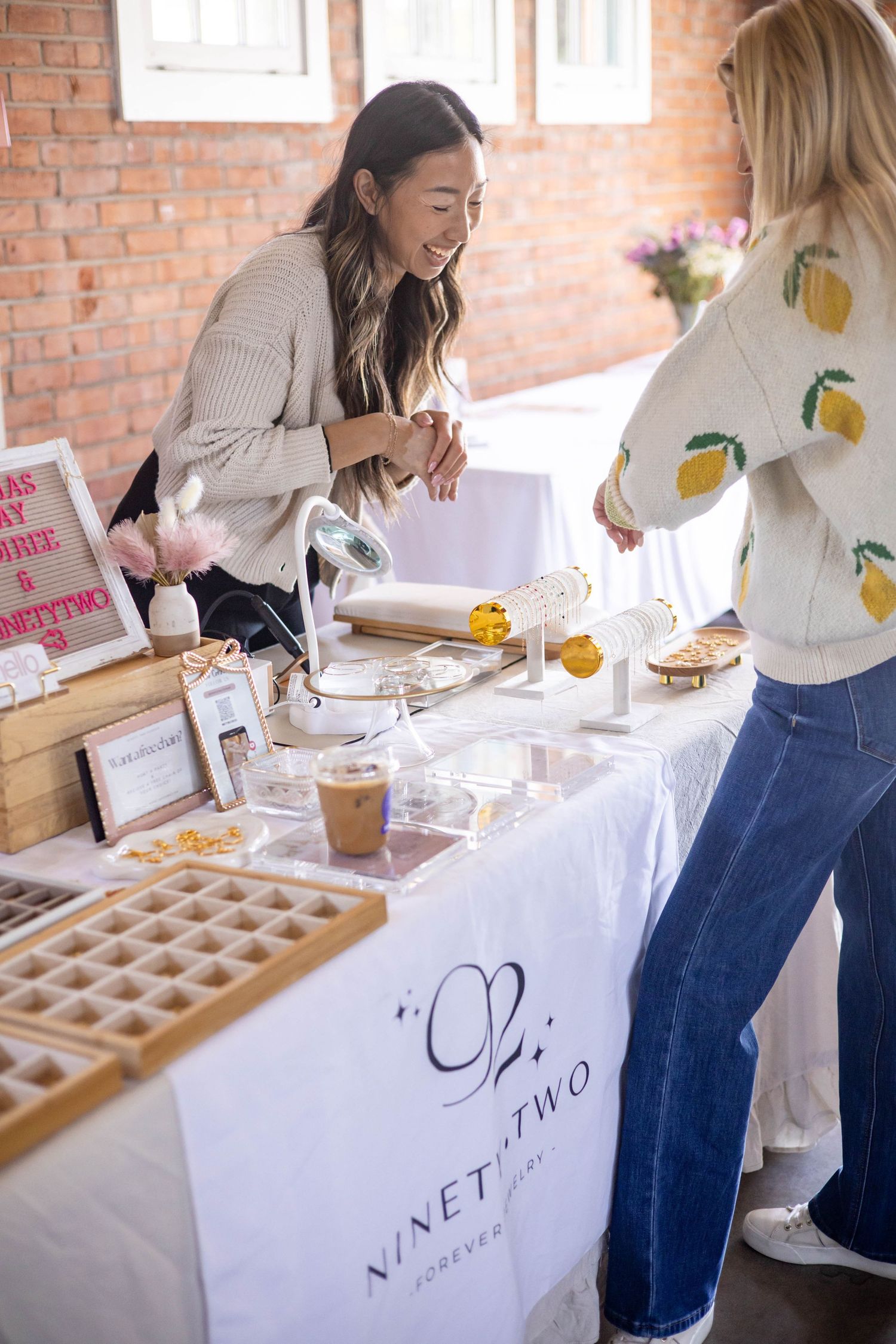 Two women at a NinetyTwo Forever vendor table displaying baked goods and treats at an indoor market event.