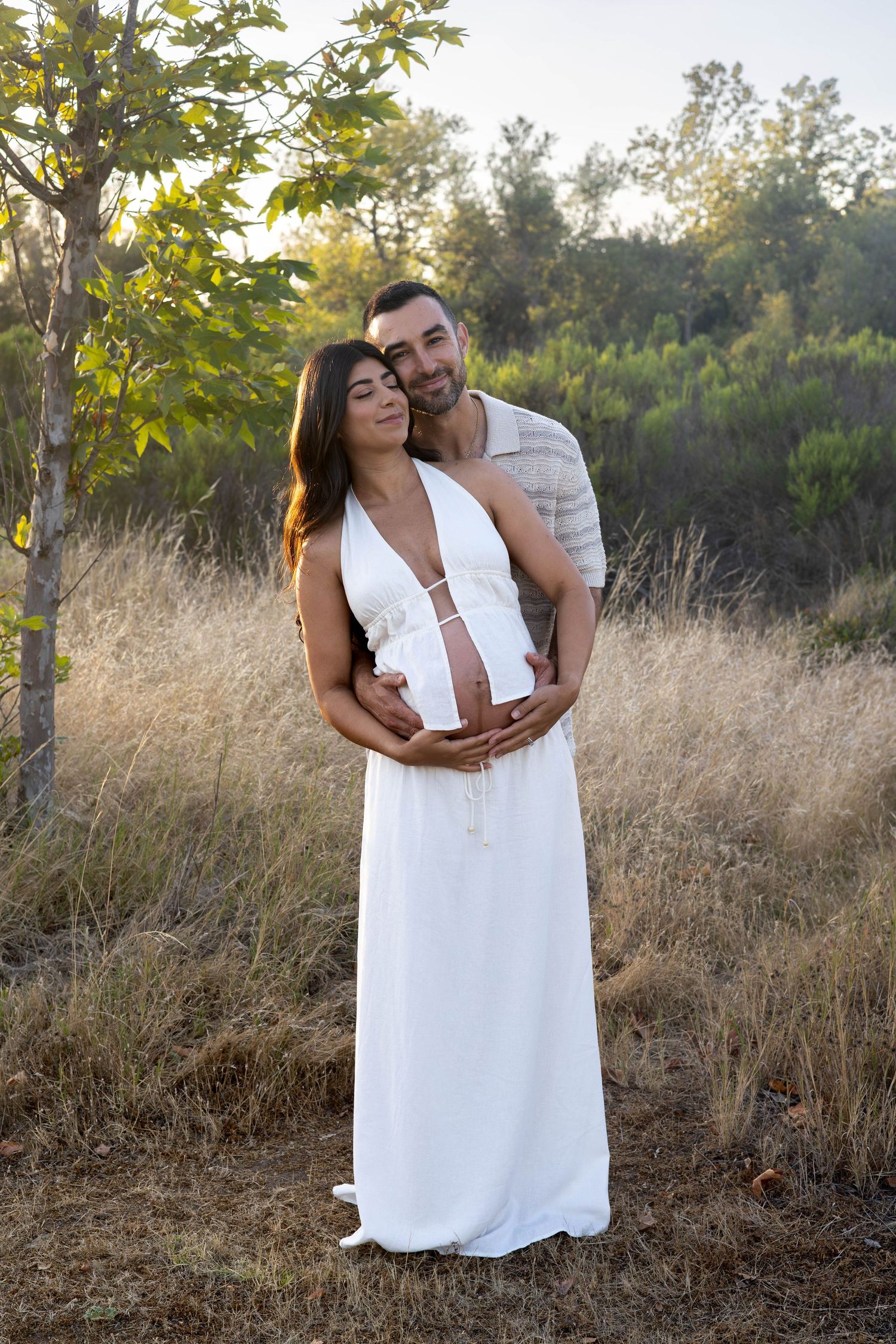Couple embracing outdoors, pregnant woman in white maxi dress, man hugging her from behind in golden field at sunset.