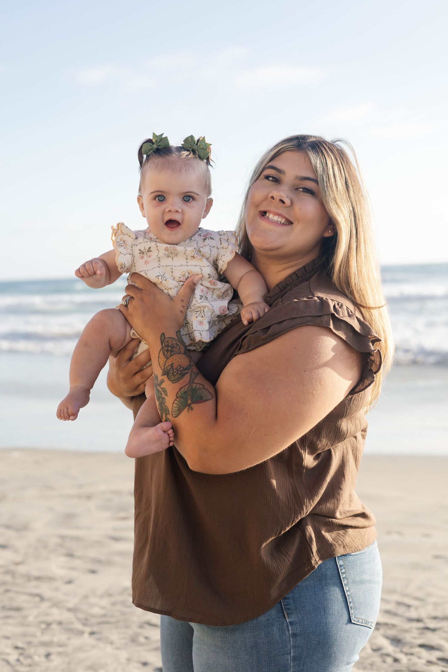 Smiling woman holding a cute baby girl on a sunny beach, wearing a brown top and jeans with a tattoo on her arm.