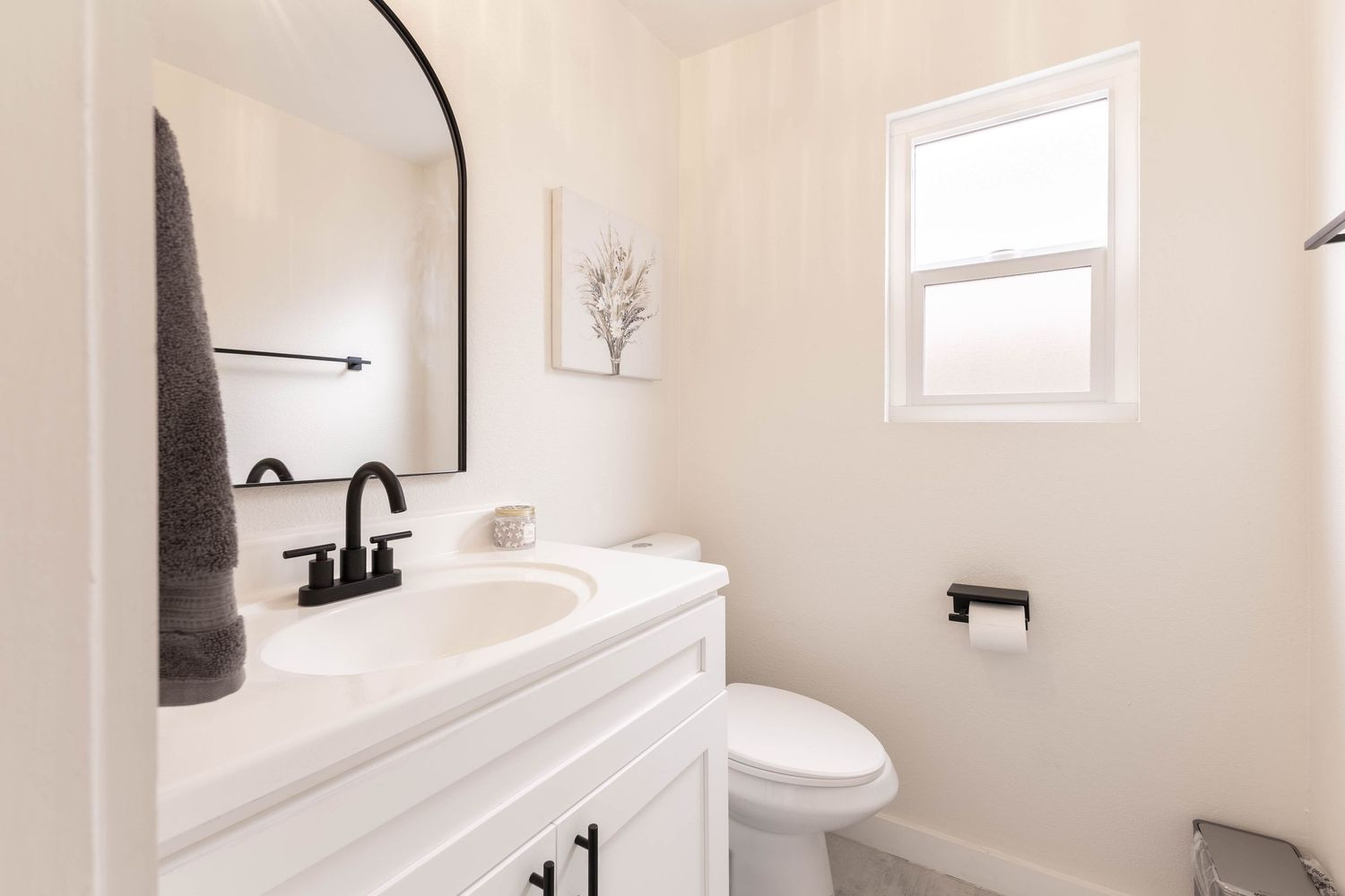 Modern white bathroom with black fixtures, vanity sink, arched mirror, and toilet near a bright window.