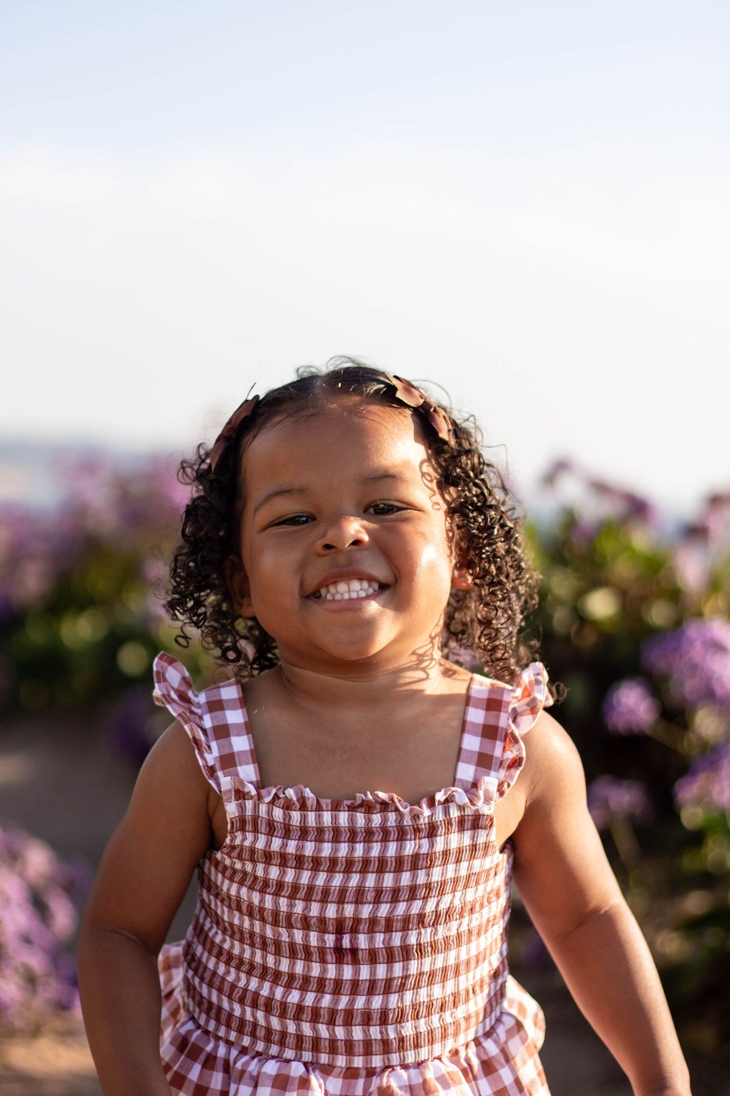 Adorable toddler girl with curly hair smiling in front of purple flowers outdoors.