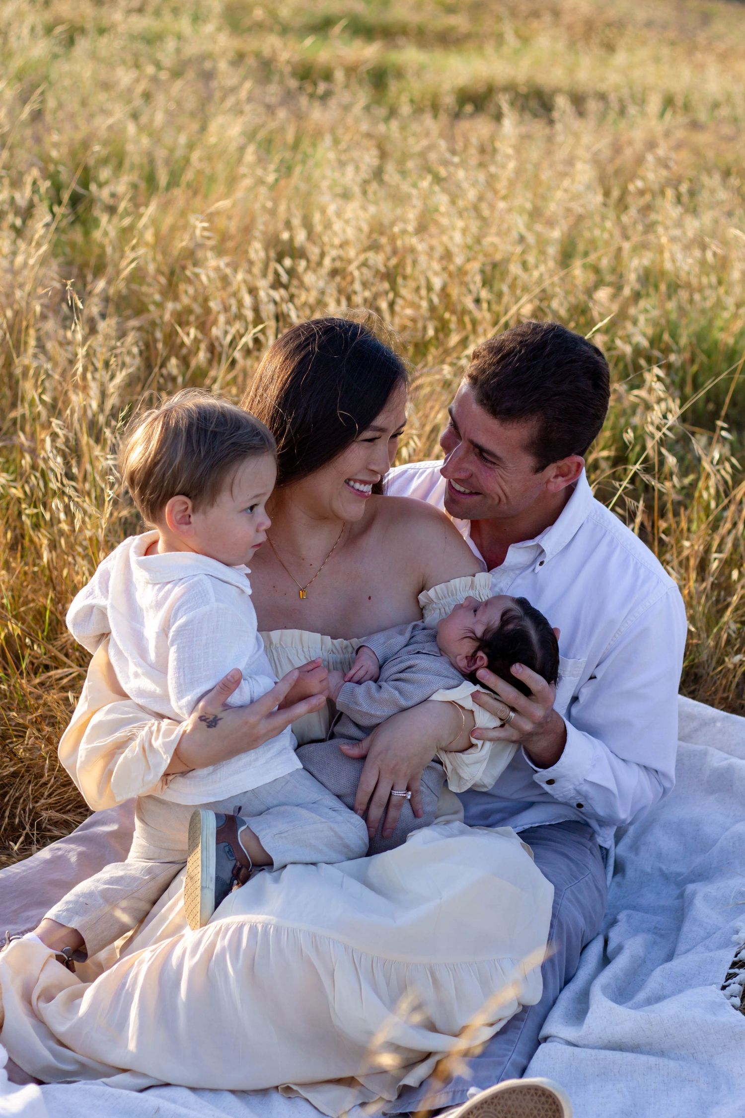 Young family sits together on blanket in golden field, parents smiling at each other with two children.