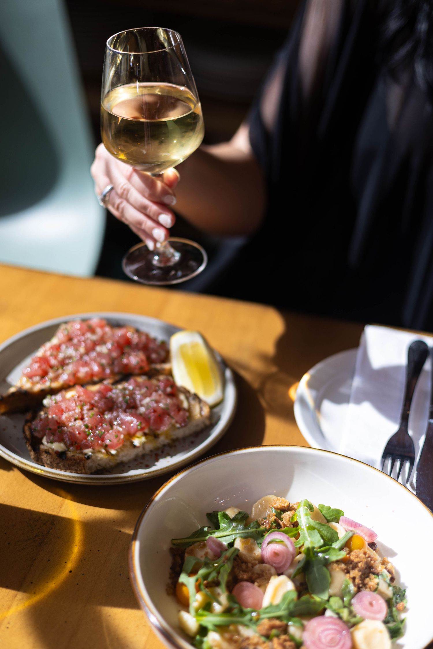 A woman holds a glass of white wine at a sunny restaurant table with bruschetta and a fresh salad.