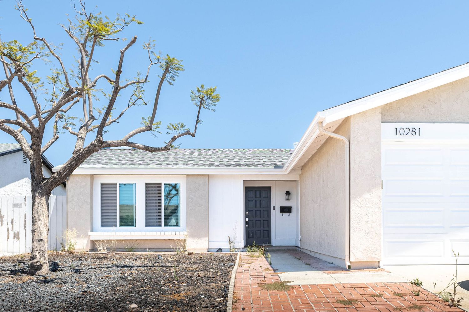 White single-story ranch home with dark front door, attached garage, and mature tree in xeriscaped front yard.