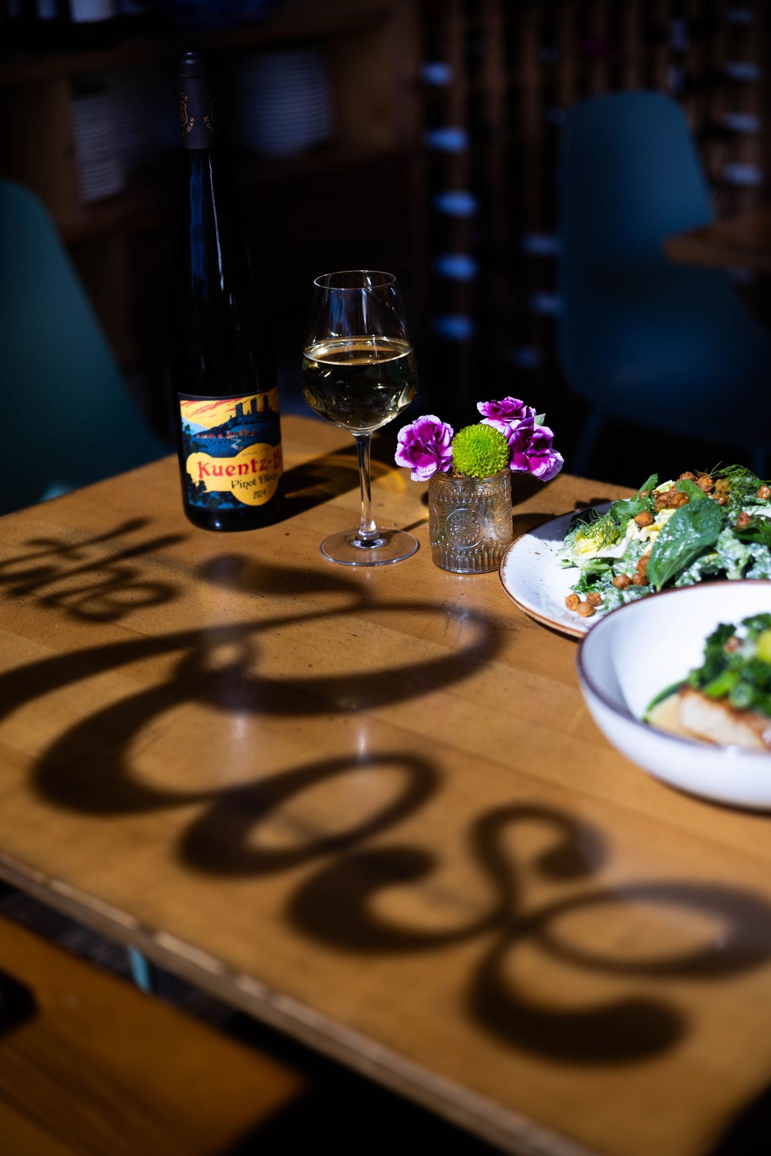 Elegant restaurant table with white wine bottle, glass, fresh salad, and decorative flowers casting artistic shadows.