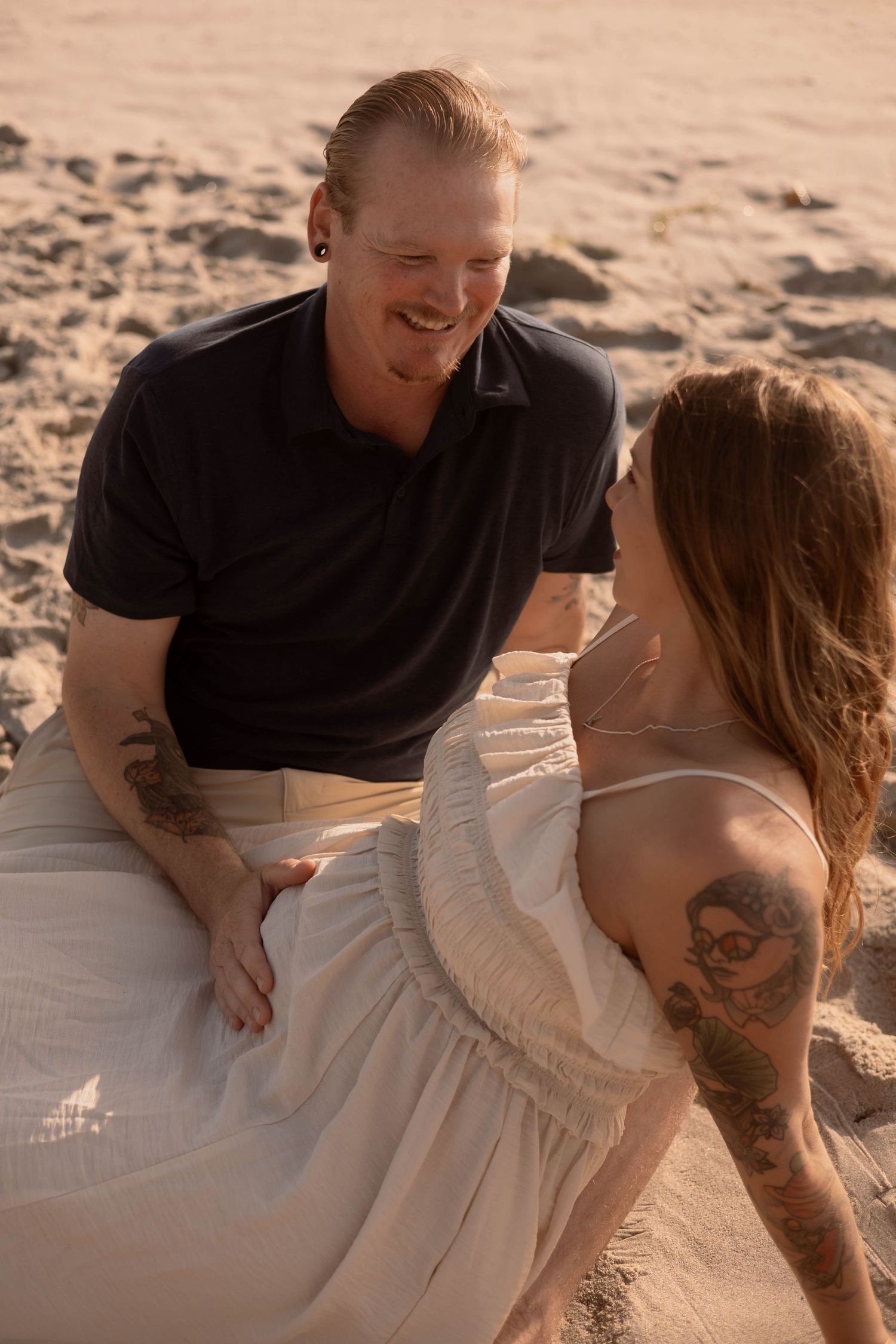Couple with tattoos sitting together on sandy beach, smiling and sharing a romantic moment at golden hour.
