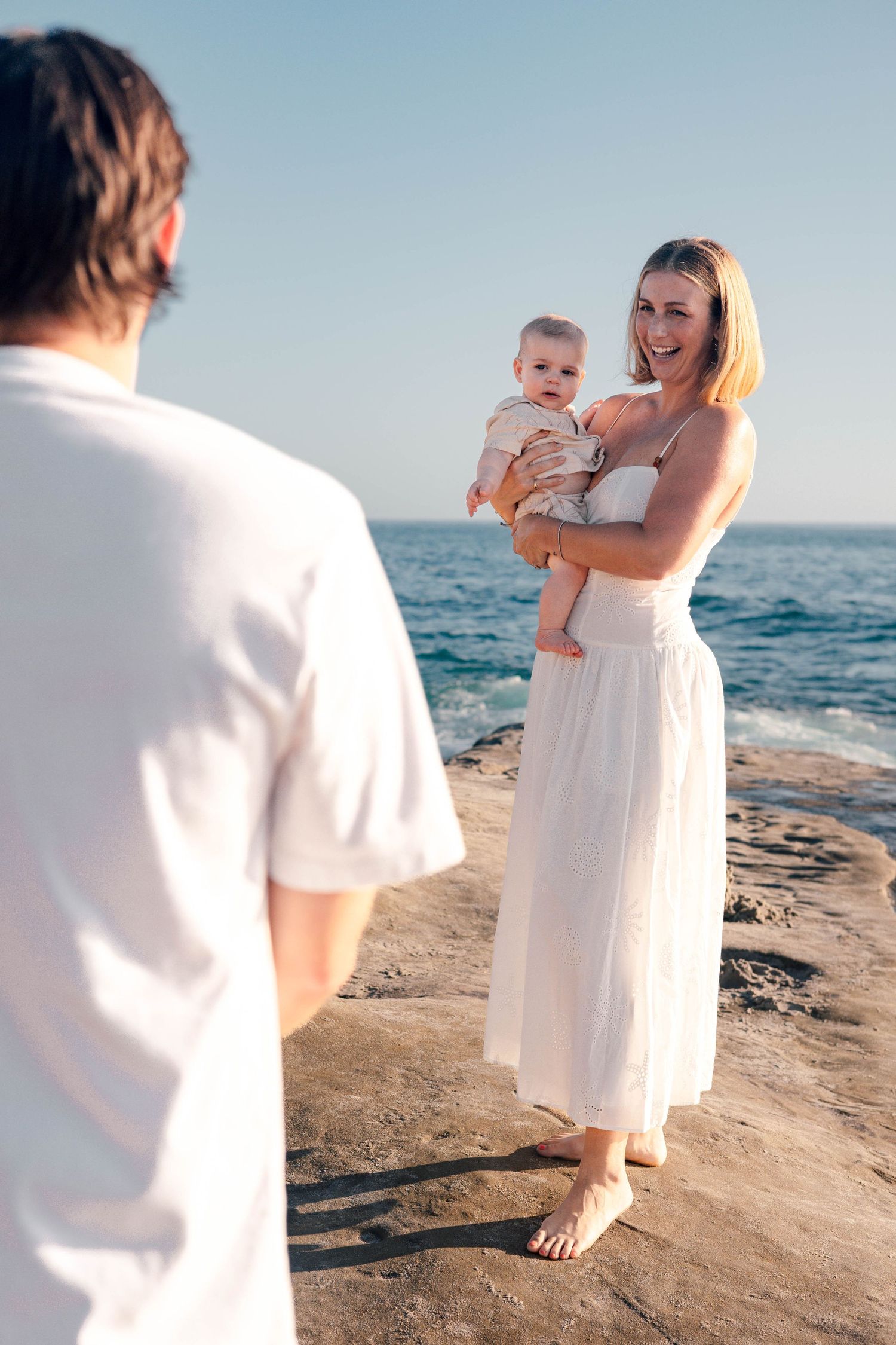 A smiling woman in white dress holds baby on rocky beach, facing a man in white shirt with ocean backdrop.