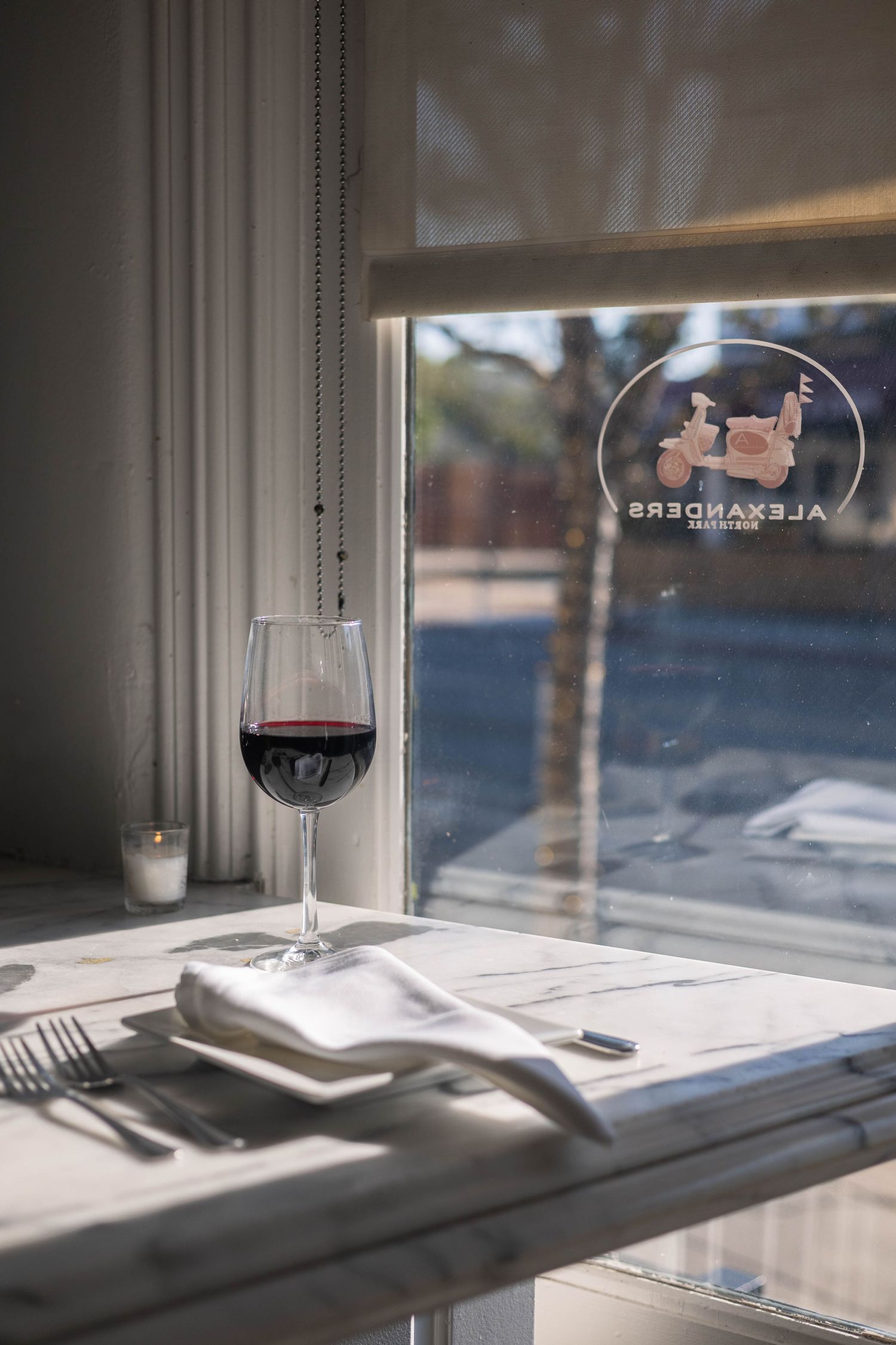 A glass of red wine on a marble table by a window at Alexander's Restaurant, with elegant place settings.