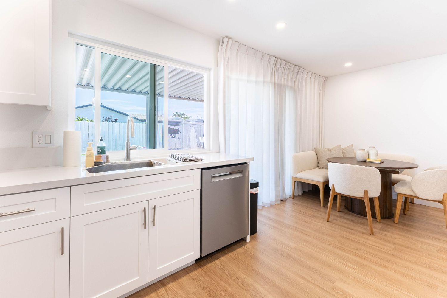 Modern white kitchen with stainless steel dishwasher, farmhouse sink, and open dining area with hardwood floors.