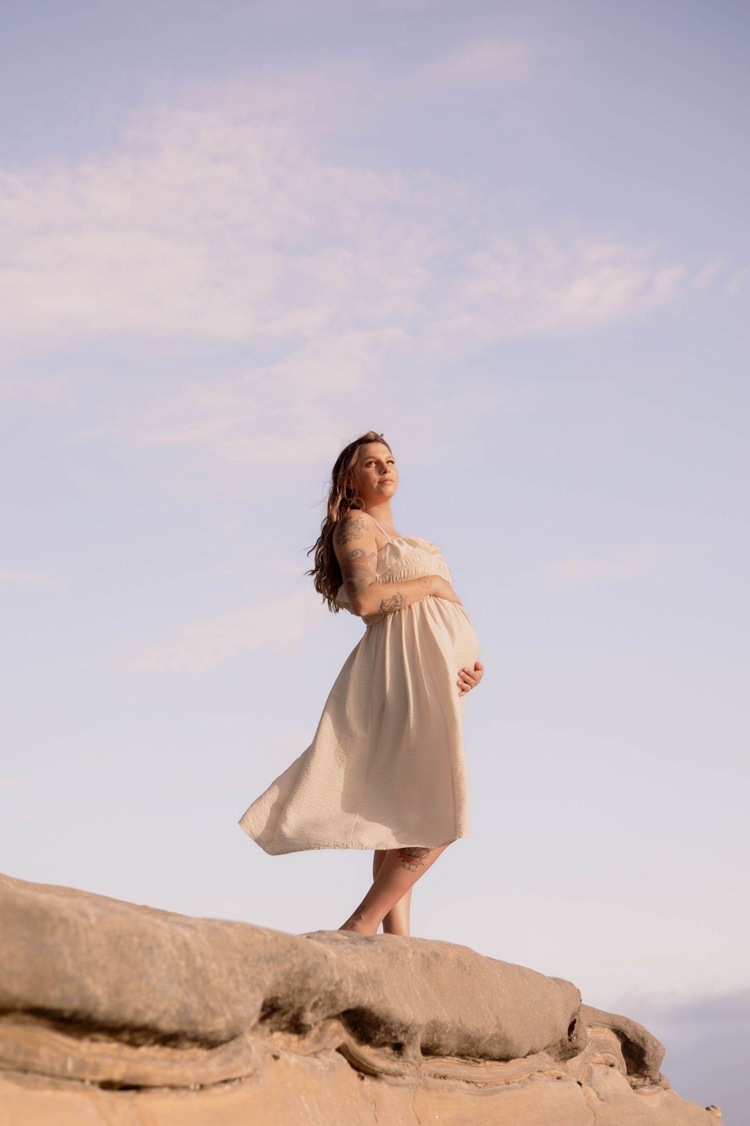 Pregnant woman in flowing white dress standing on desert rocks against a pastel blue sky.