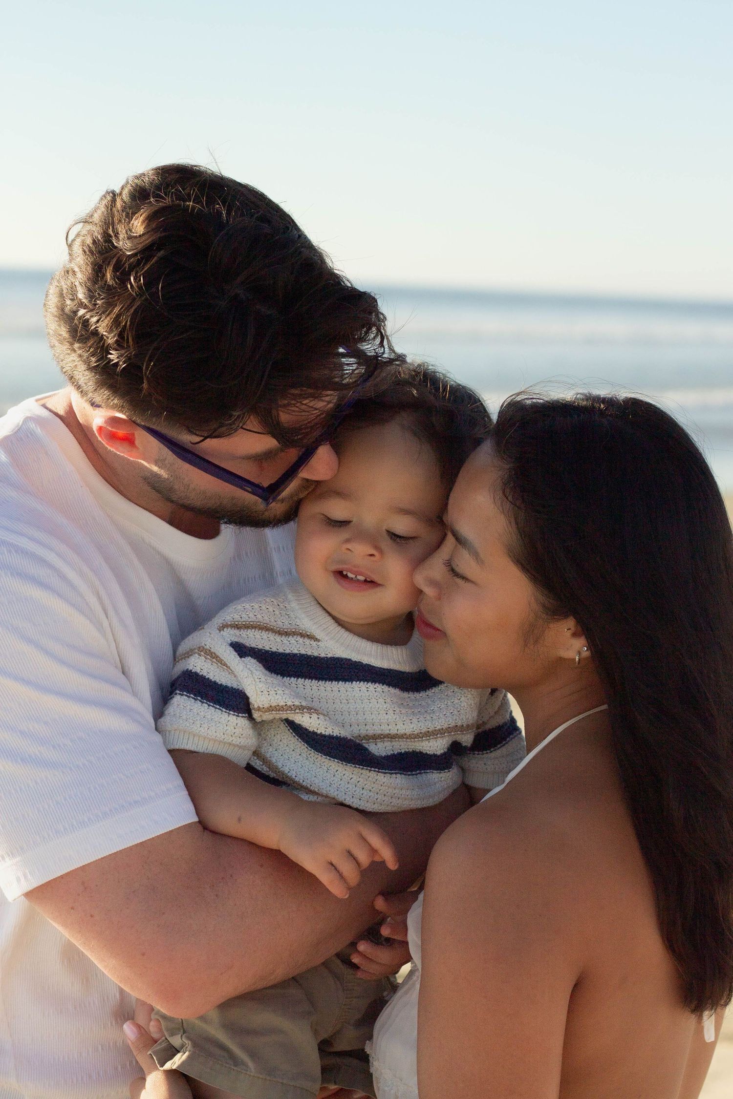A loving family shares a tender moment as parents kiss their smiling baby at the beach.