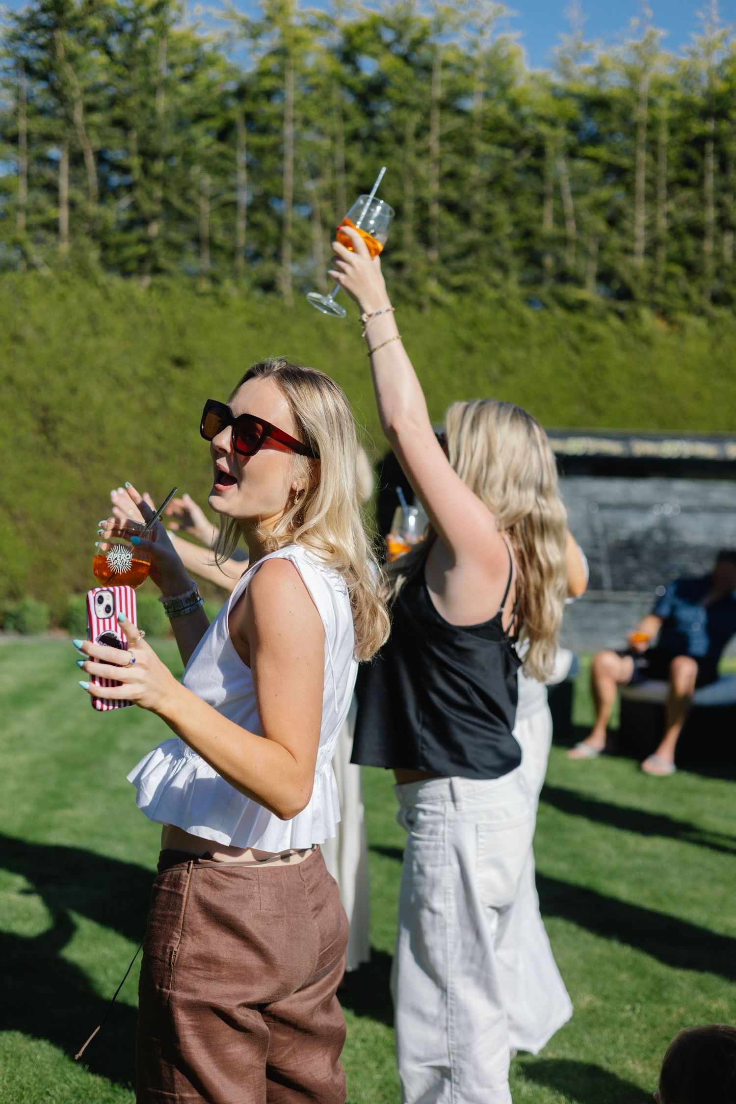 Two blonde women dancing outdoors on green grass, holding colorful drinks, wearing casual summer outfits on a sunny day.