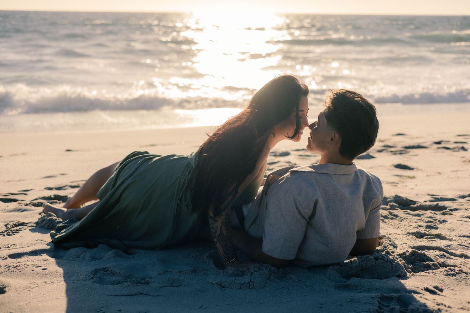 Romantic couple lying on sandy beach at sunset, sharing an intimate moment with ocean waves in background.
