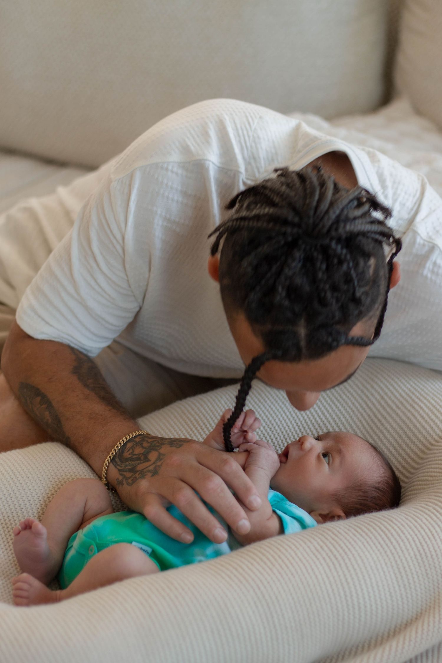 Tattooed father with dreadlocks tenderly bonding with his newborn baby in a teal onesie on a couch.