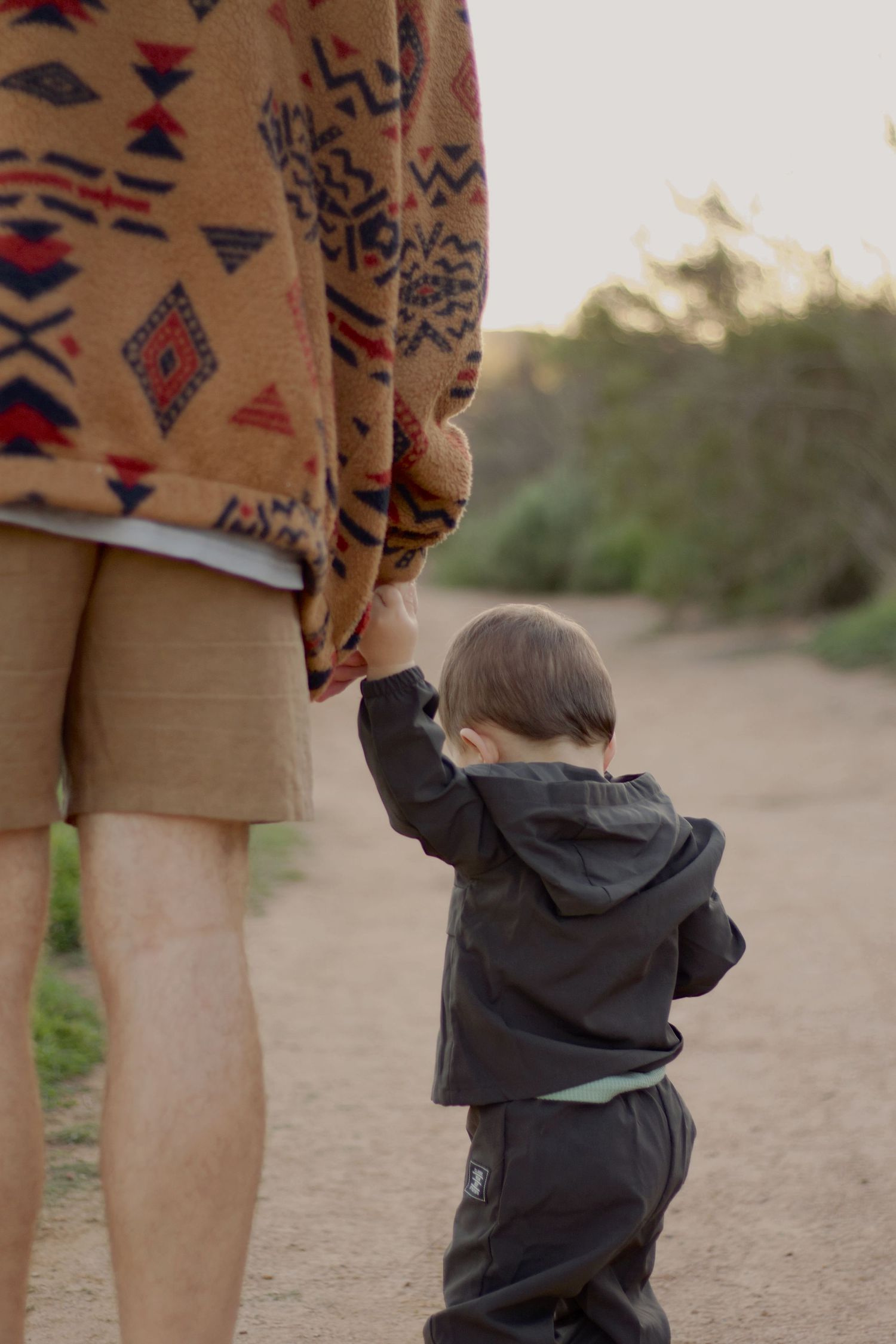 A toddler in a dark jacket holds an adult's hand while walking on a dirt path at sunset.