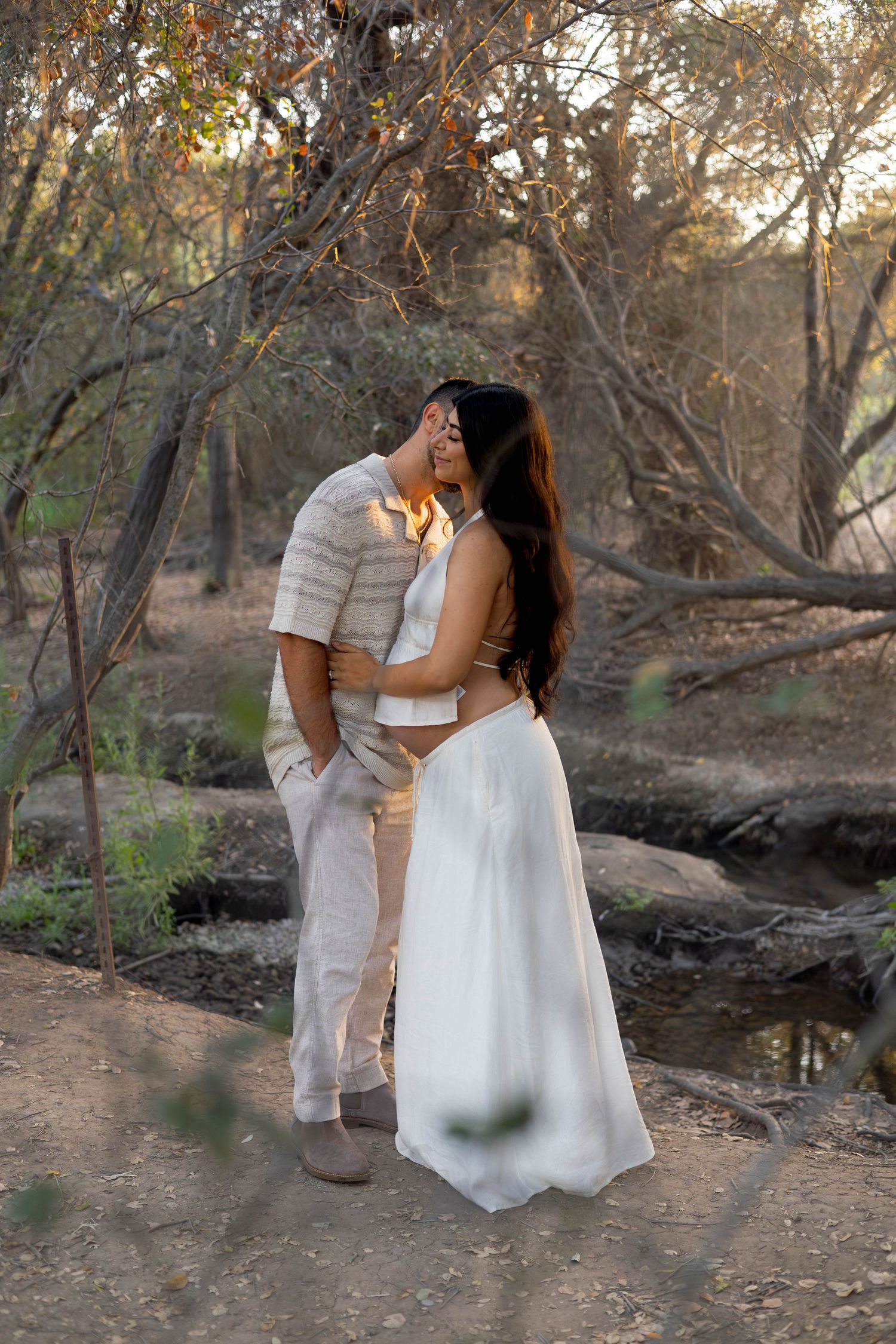 Couple sharing a romantic kiss outdoors in a wooded setting, woman in white dress, man in beige outfit.