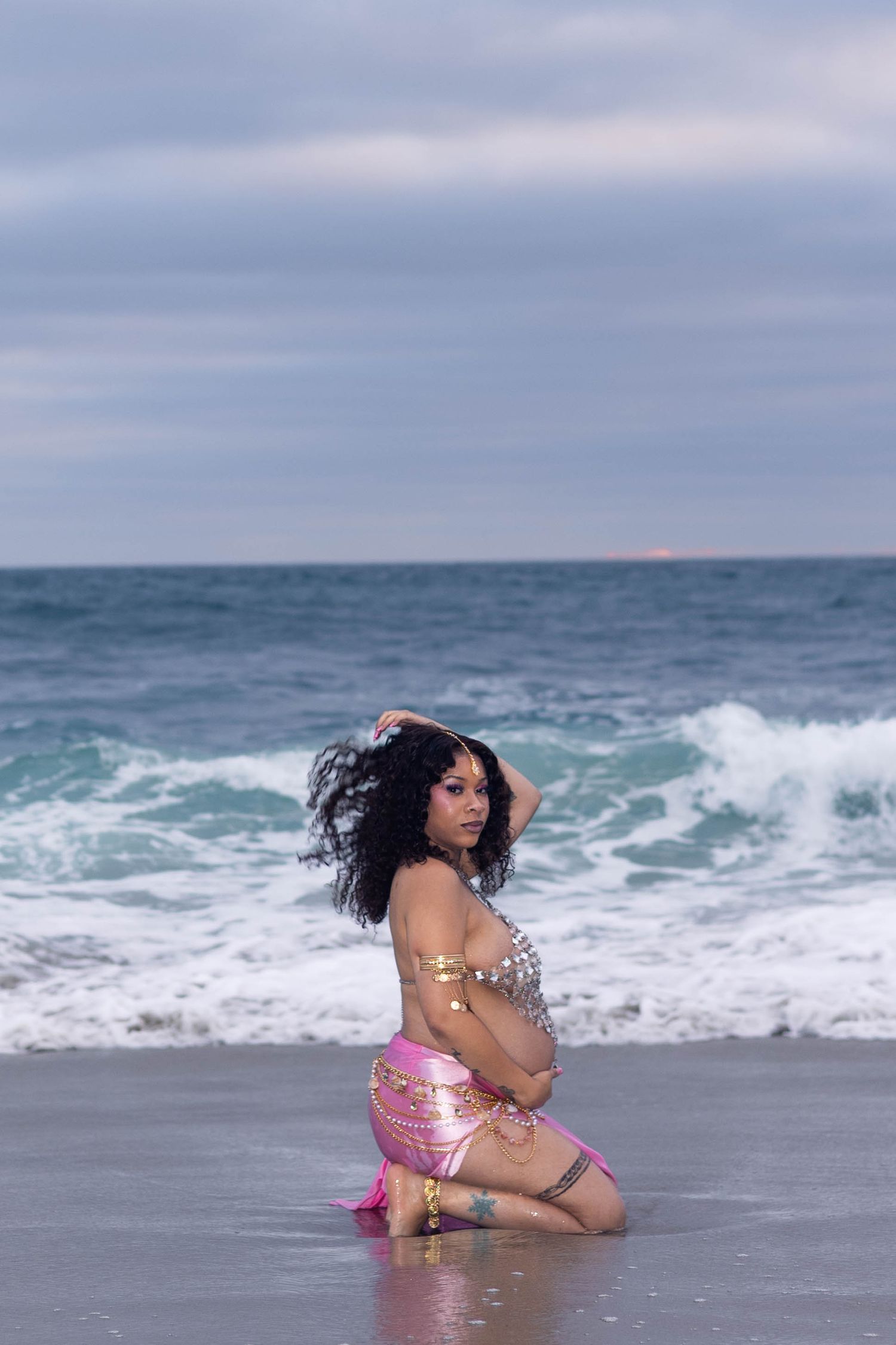 Pregnant woman in pink and gold outfit kneeling on beach with ocean waves behind her.