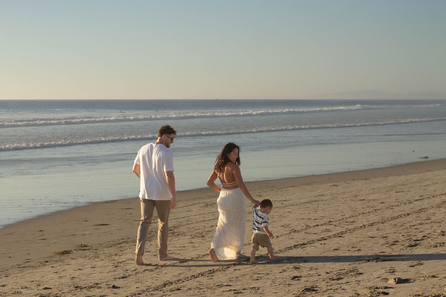 A family strolls along the shoreline as parents guide their toddler on a sunny beach.
