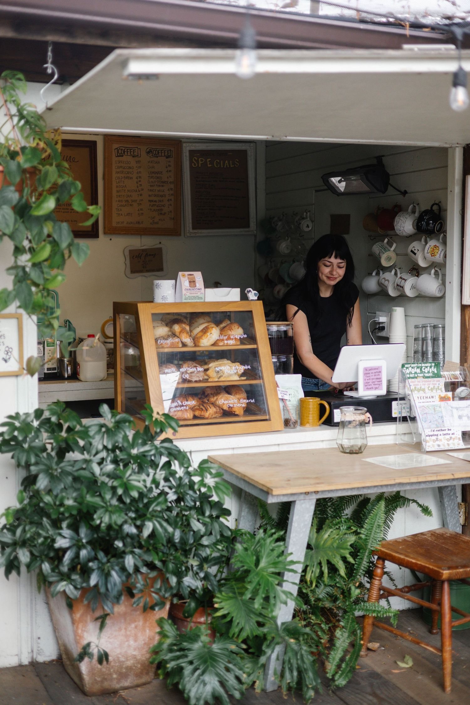 A smiling woman works behind a charming outdoor bakery counter with pastries on display, surrounded by lush green plants.