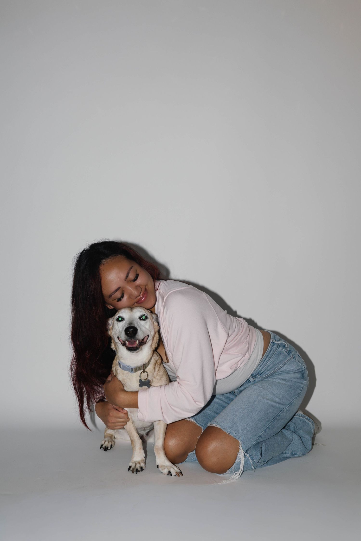 Young woman in pink top and jeans kneeling on floor hugging a happy white dog in a studio setting.