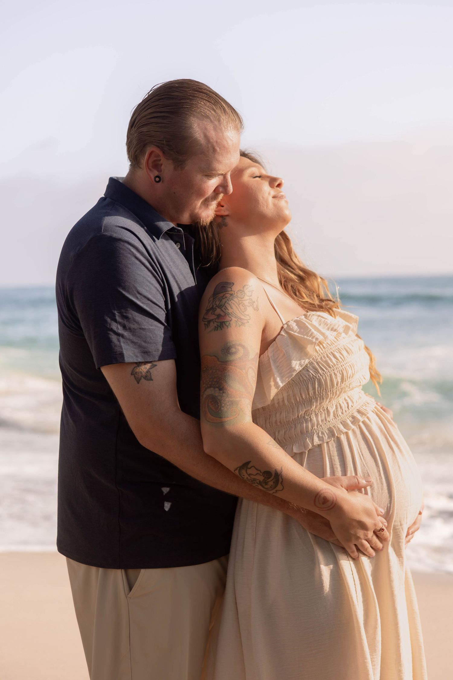 A tattooed couple embraces on a beach, the pregnant woman leaning back as her partner holds her bump near ocean waves.