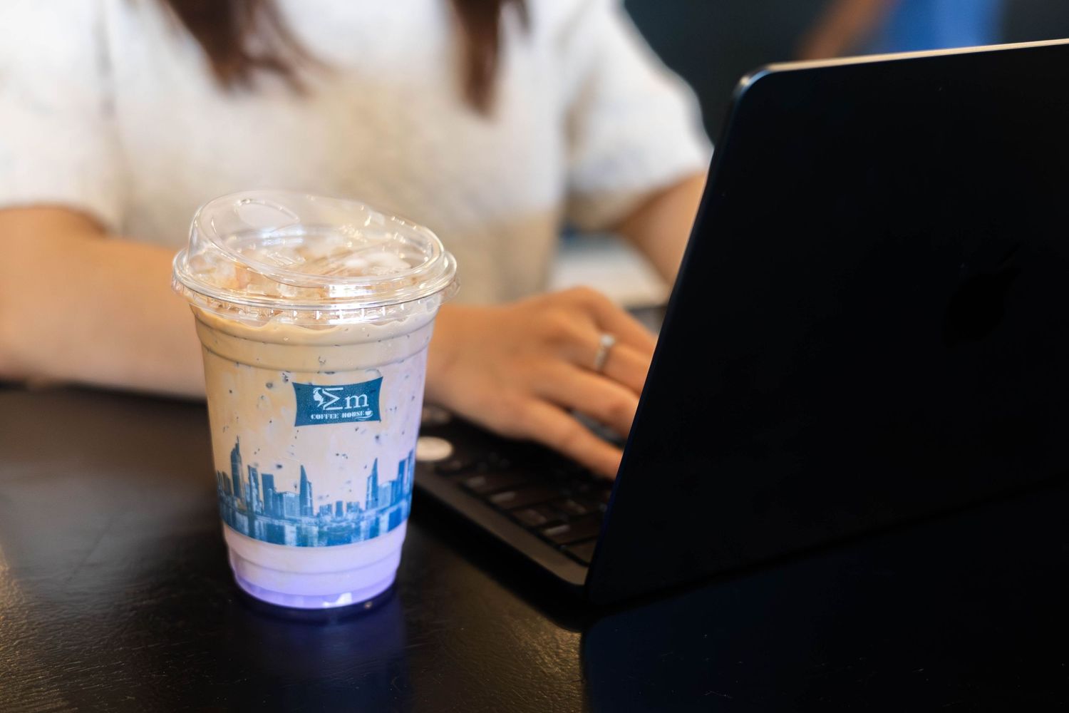 Person working on laptop beside an iced coffee drink in a branded plastic cup on a dark table.