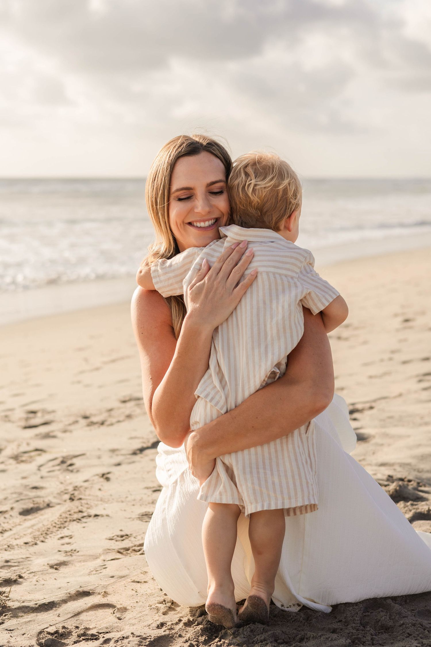 A smiling mother hugs her toddler on a sandy beach at sunset, both wearing light neutral outfits.
