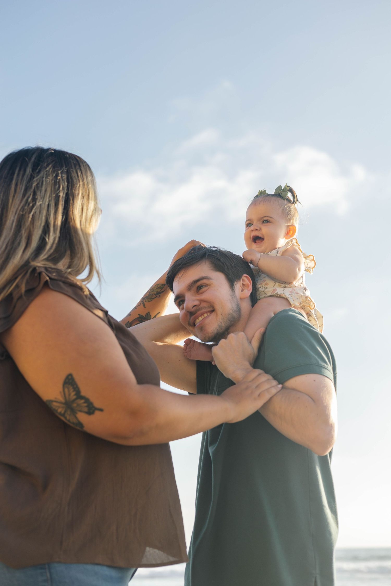 A happy family at the beach, with a smiling dad holding a laughing baby girl while mom lovingly supports them.