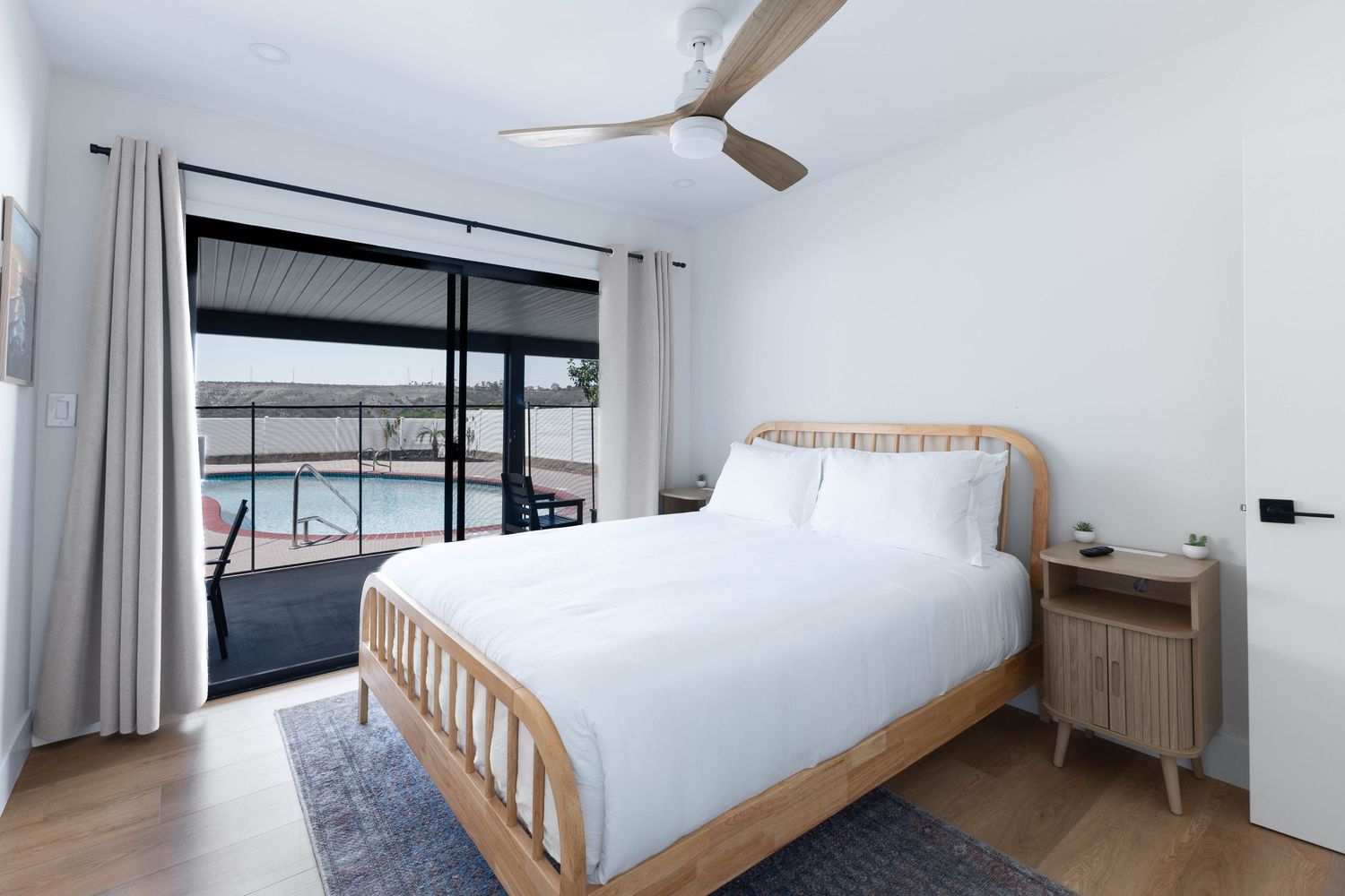 Modern bedroom with rattan bed, white bedding, hardwood floors, and sliding glass doors opening to a pool view.
