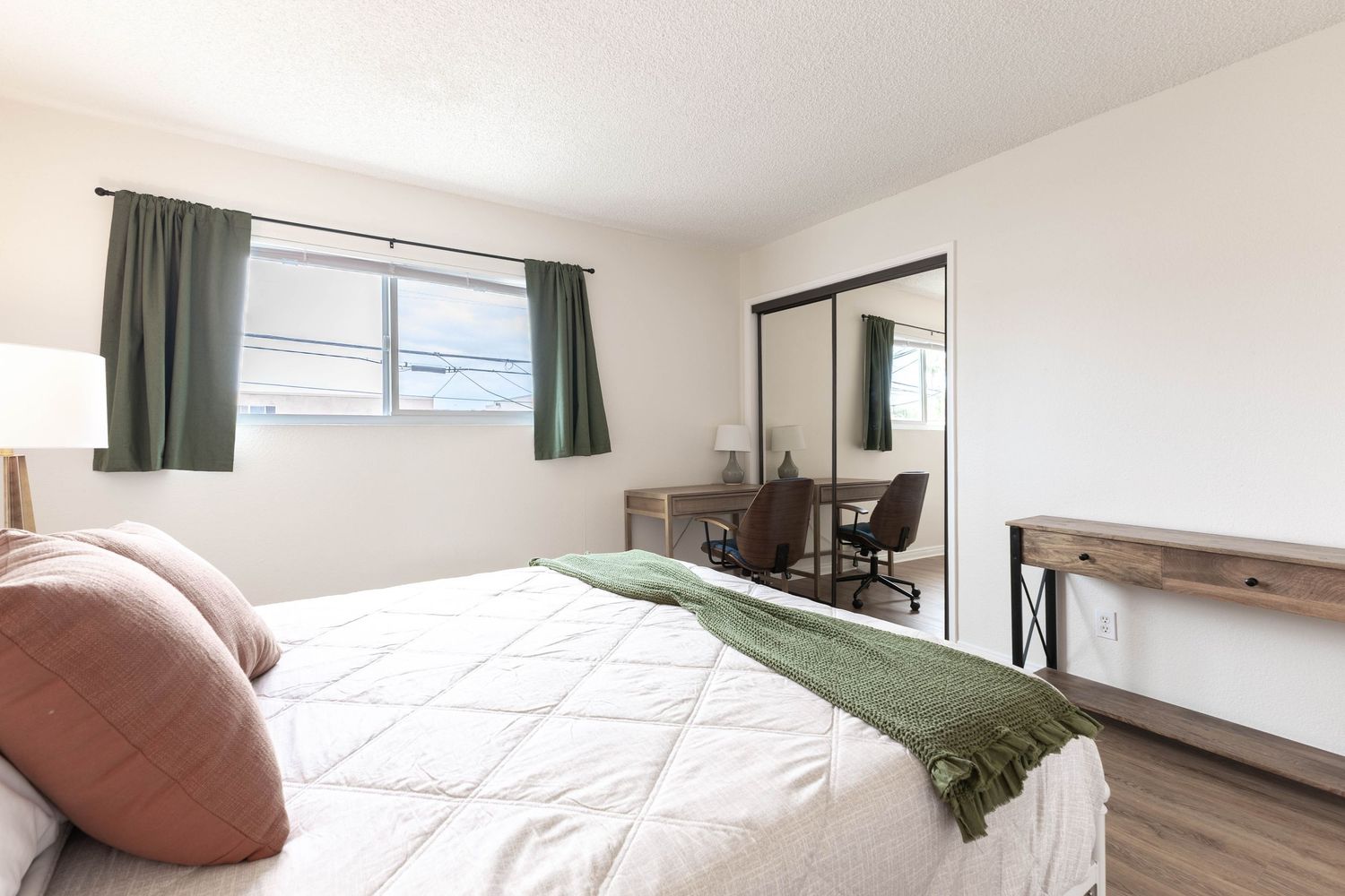 Bright bedroom with a queen bed, green curtains, mirrored closet, and a wooden desk in a clean modern space.