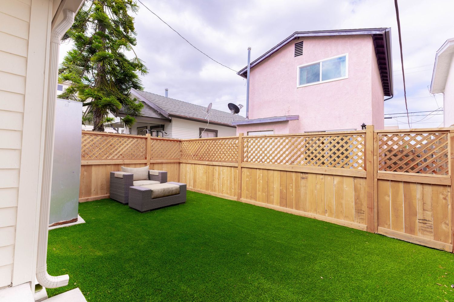 Backyard with lush artificial grass, wooden lattice privacy fence, and outdoor seating area under cloudy skies.