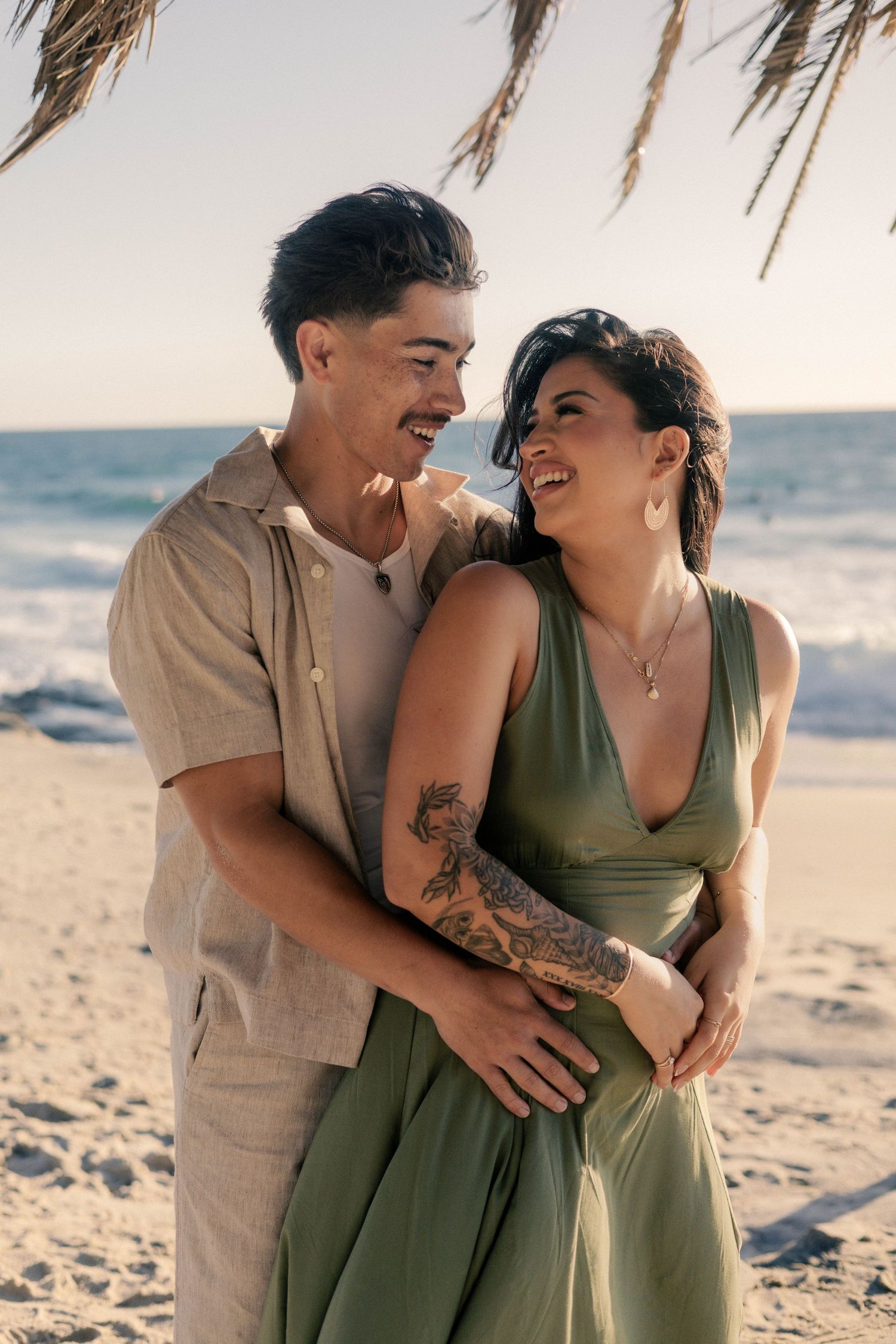 Laughing couple embracing on sandy beach at sunset, woman with tattoos in green dress held by partner.