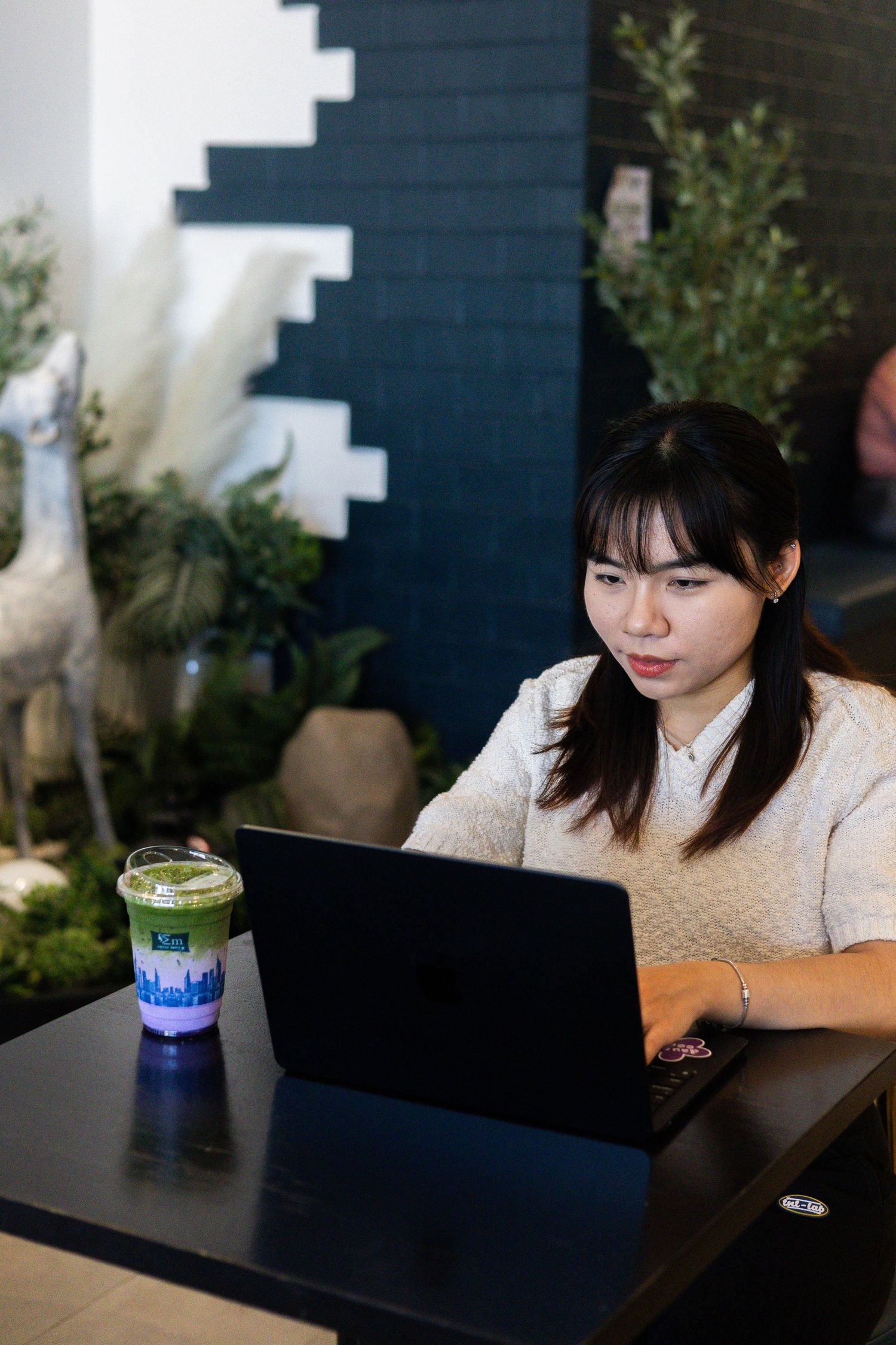 Young Asian woman working on laptop at cafe table with matcha drink, surrounded by green plants and decorative deer statue.