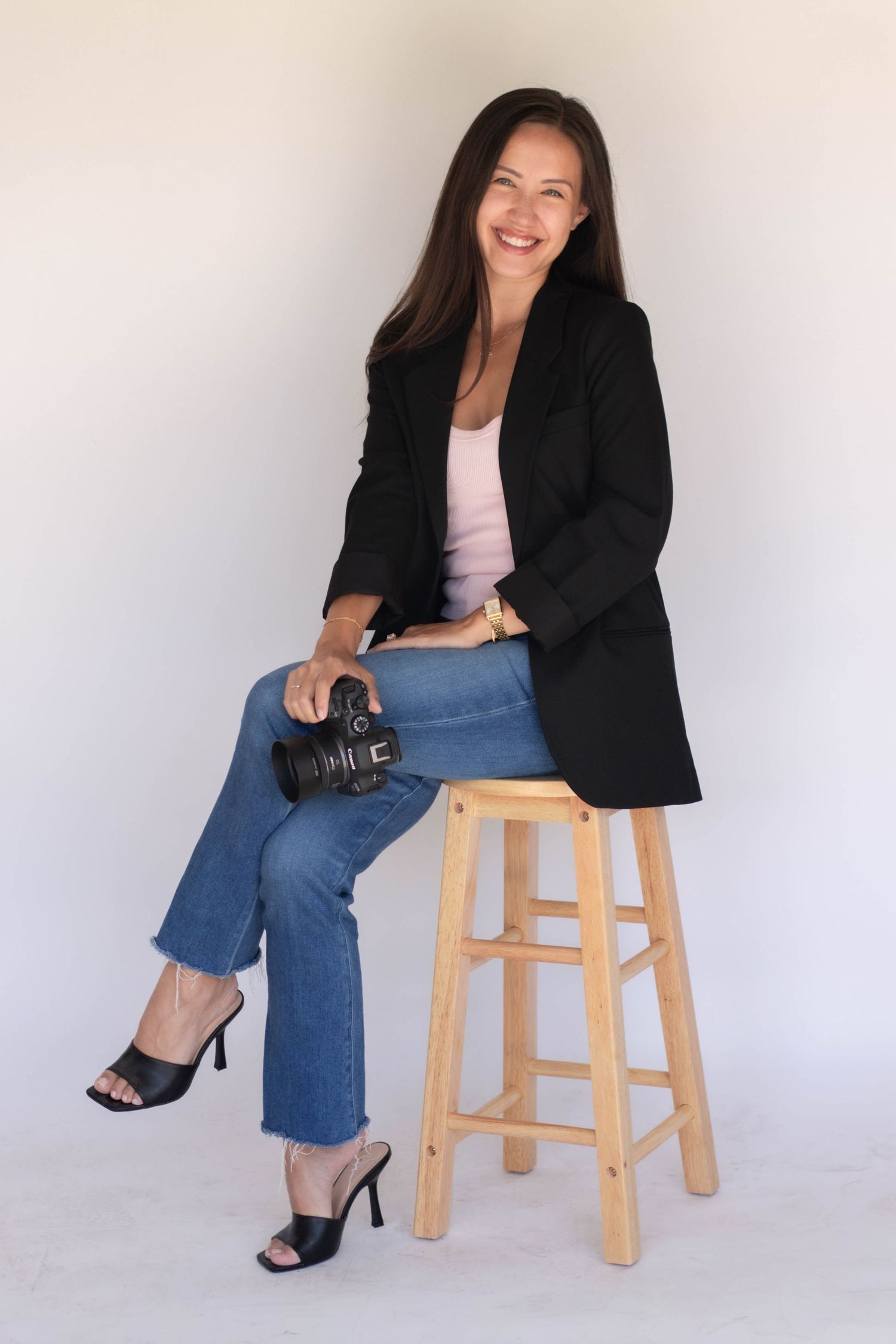 Smiling woman in black blazer and jeans sits on wooden stool holding camera, wearing heels against white background.