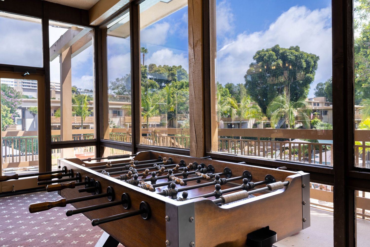 Foosball table in a bright sunlit game room with large windows overlooking lush green trees and an outdoor patio area.
