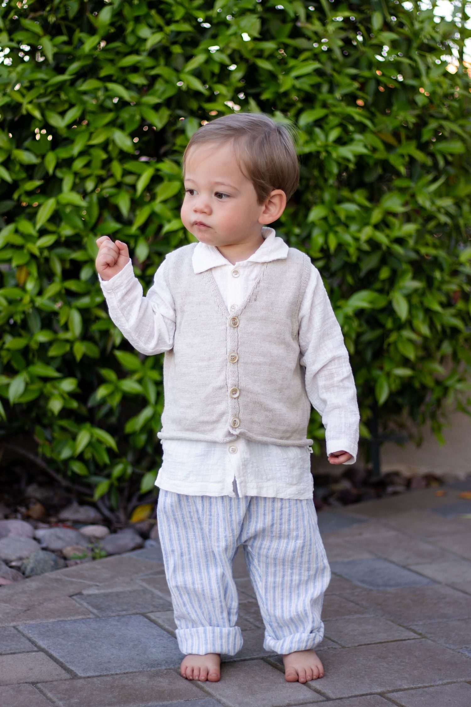 An adorable toddler in a linen vest and striped pants stands barefoot on a garden patio.