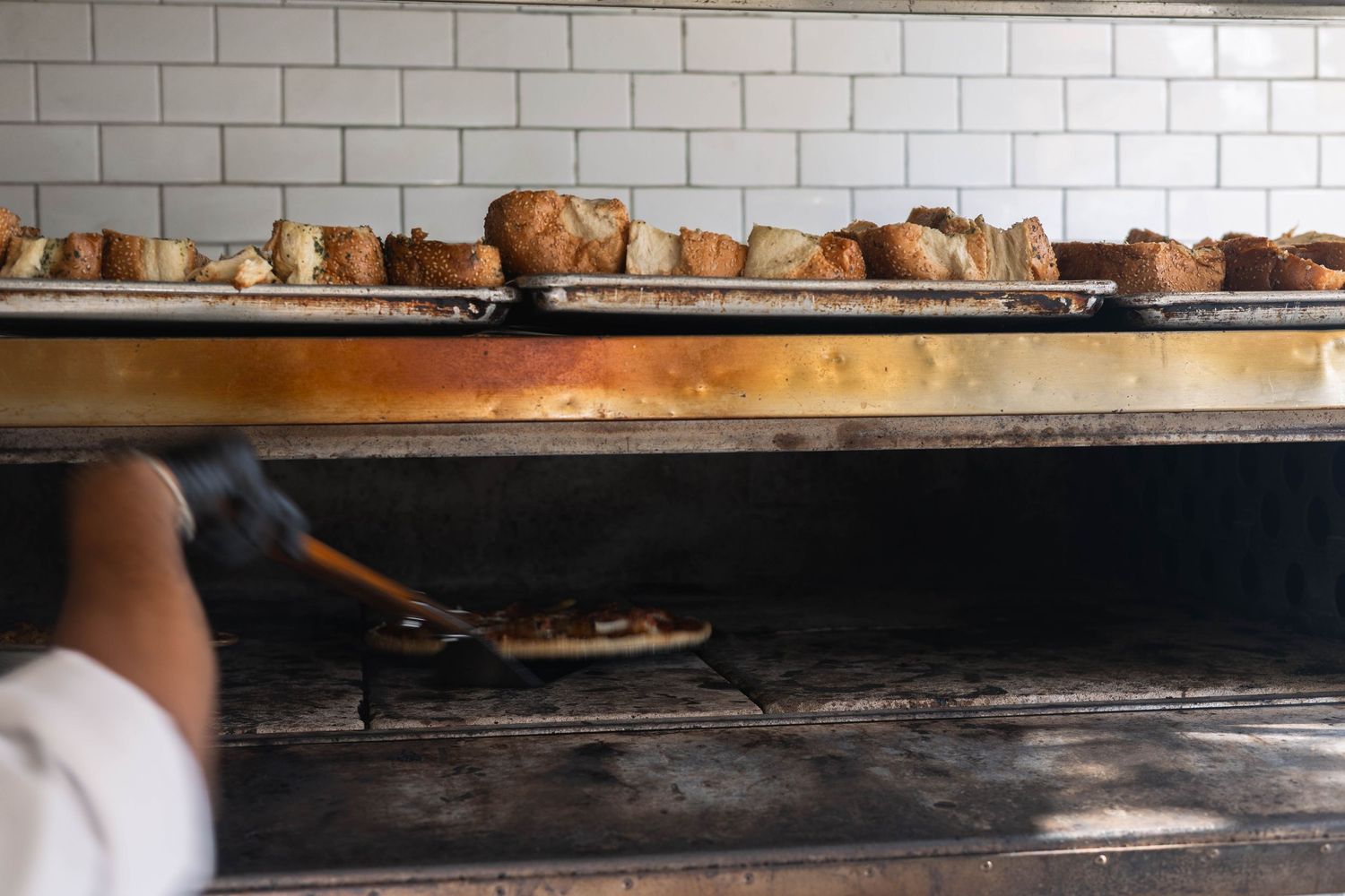 Baker sliding pizza into a wood-fired oven with fresh baked breads on a rack above the flames.